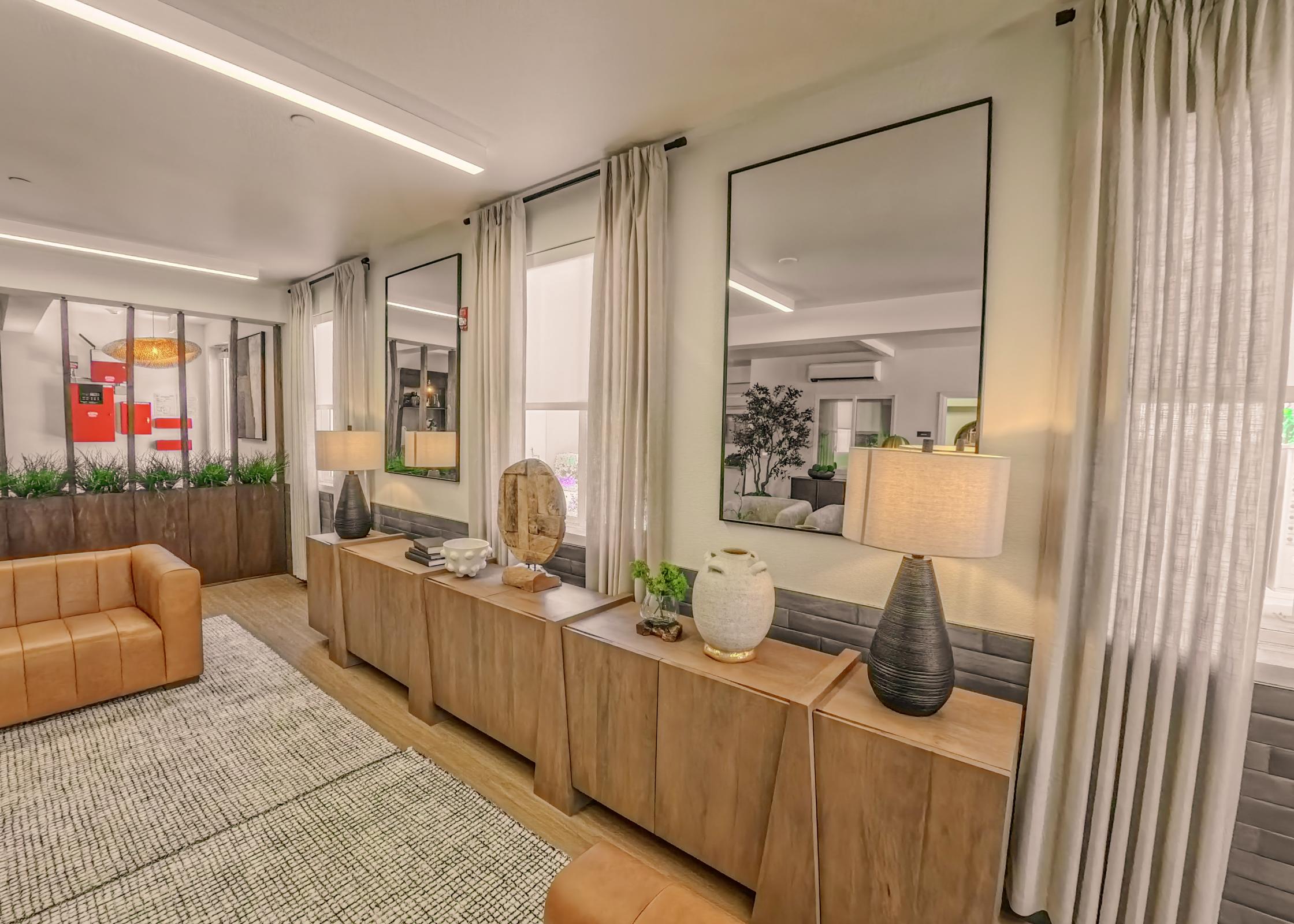 A modern living room featuring a leather sofa, wooden sideboard with decorative items, table lamps, and large mirrors. Natural light filters through sheer curtains, illuminating plants in the background and creating a cozy, inviting atmosphere.