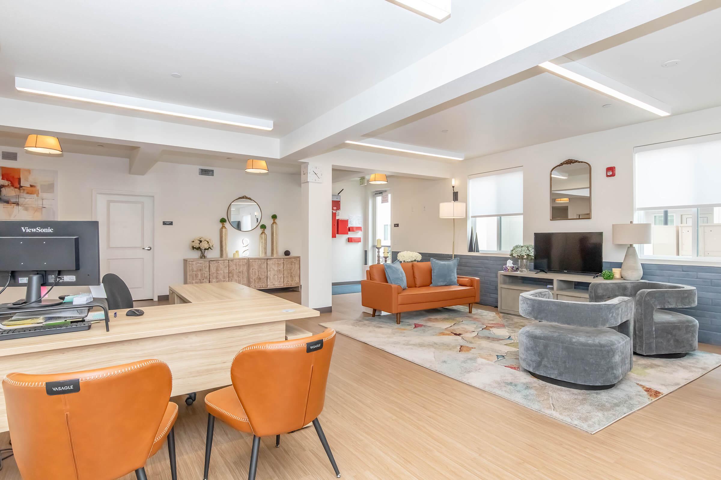 A modern communal living area featuring a wooden table with office chairs, a cozy sitting area with an orange sofa and gray chairs, a rug with a geometric pattern, a television, decorative plants, and stylish lighting, all in a bright and inviting atmosphere.