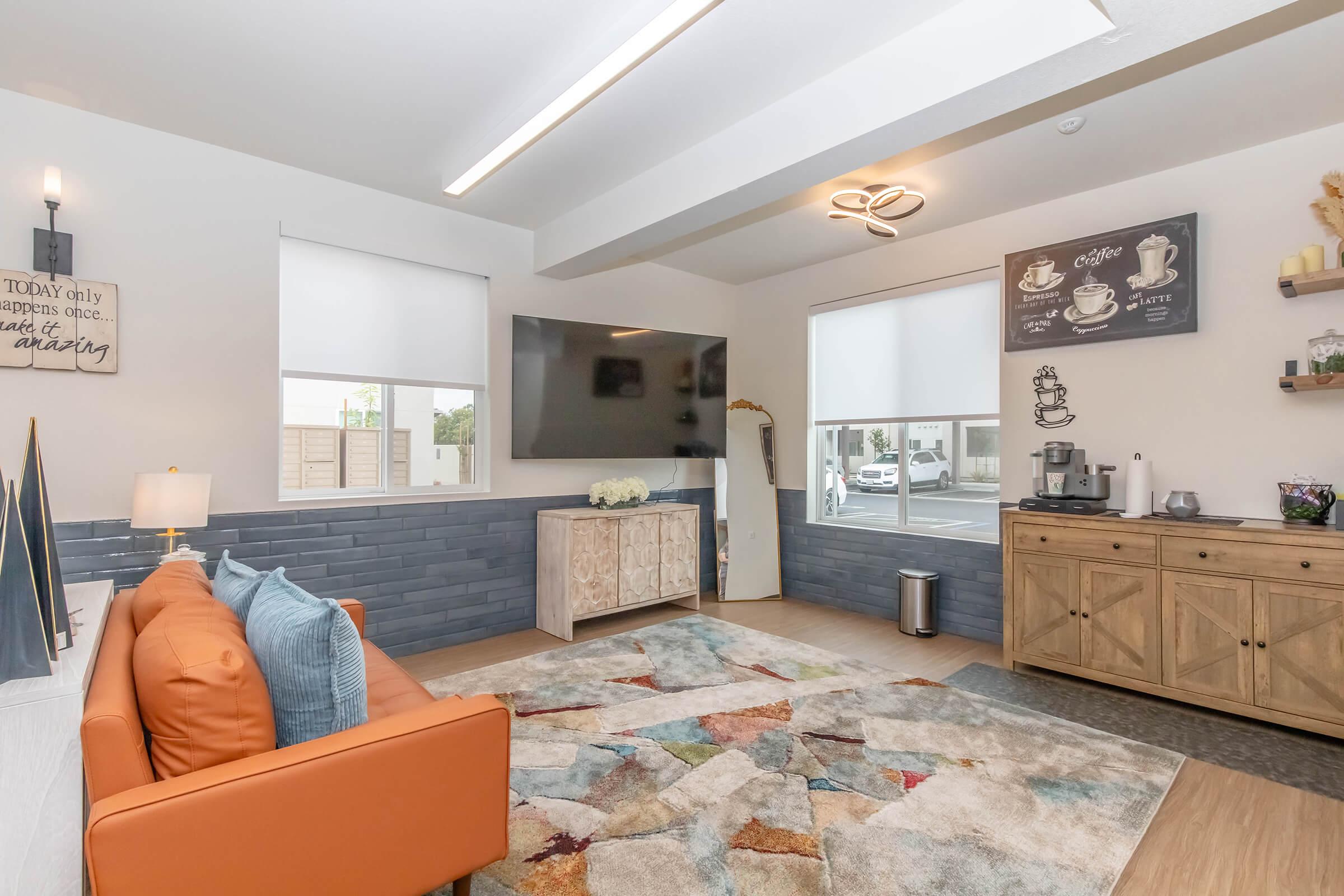 A modern living space featuring a cozy orange sofa, a large TV mounted on the wall, and a stylish rug with various colors. Windows allow natural light, and there’s a wooden cabinet with decorative items and a coffee station. Wall art includes coffee-themed décor.