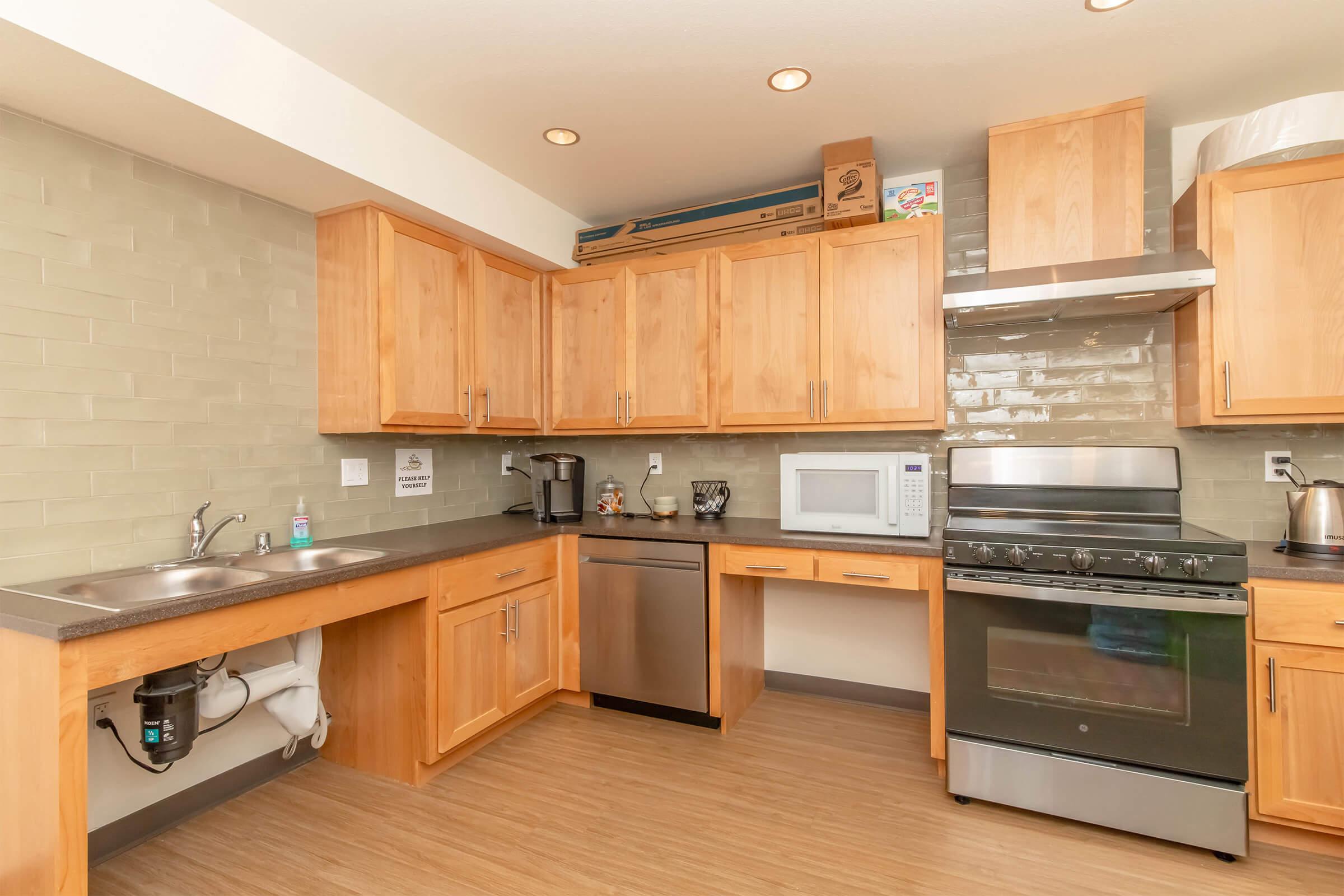 Modern kitchen featuring wooden cabinets, a stainless steel dishwasher and oven, a microwave, and a double sink. The countertop is dark, and the backsplash consists of light-colored tiles. Natural light illuminates the space, creating a warm and inviting atmosphere.