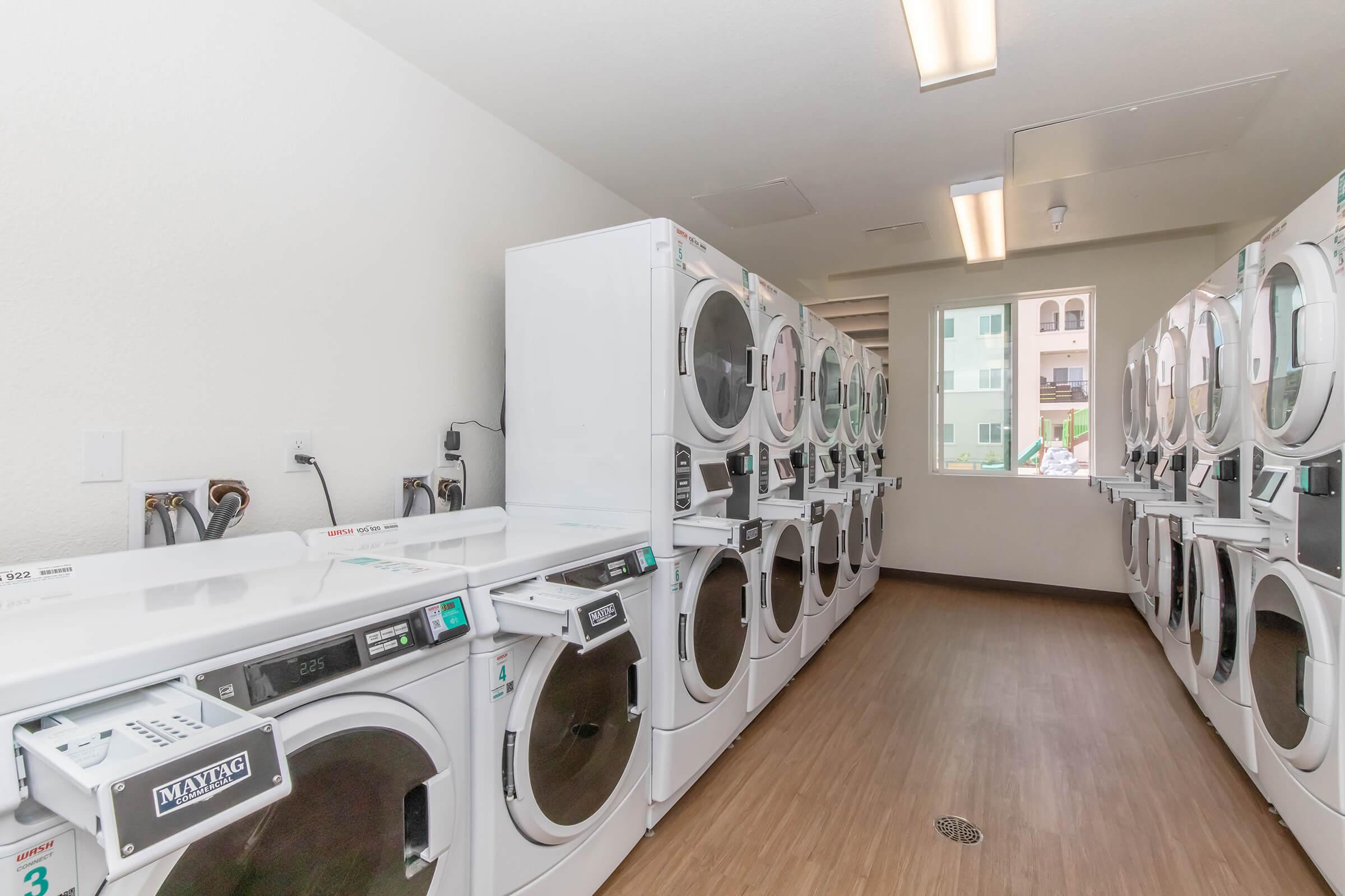 A laundry room featuring several rows of white washing machines and dryers lined against the walls. The space has a clean, modern design with wooden flooring and large windows allowing natural light. There are utility sinks and shelves for laundry supplies visible.