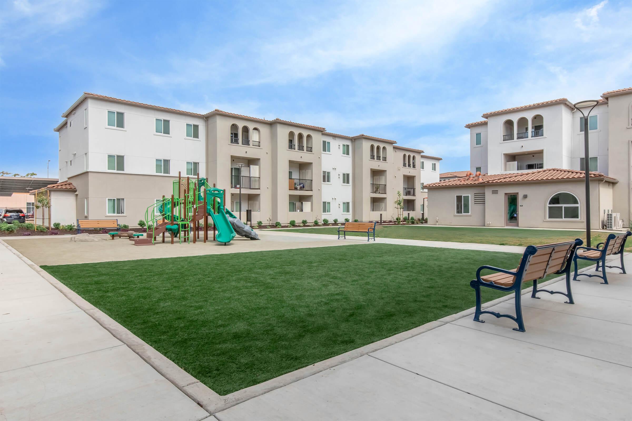 A landscaped courtyard featuring green grass, a playground with slides and climbing structures, and several benches. Surrounding the area are modern multi-story residential buildings with beige and white exteriors. The sky is clear, creating a bright environment.