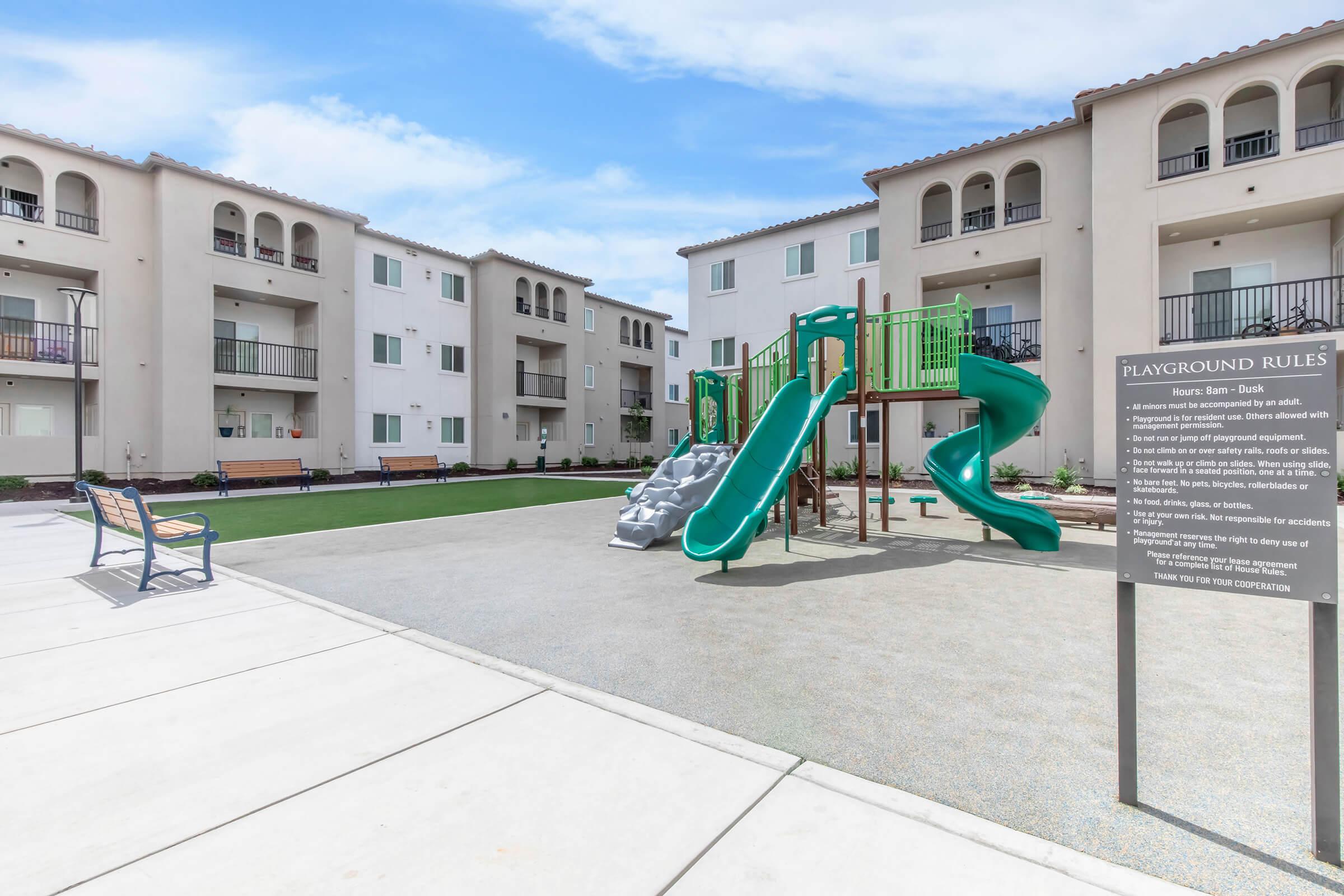 A playground area featuring a green slide and climbing structure, surrounded by new apartment buildings. A sign with playground rules stands nearby, and benches are placed along the path. The sky is clear with a few clouds, creating a bright and inviting atmosphere.