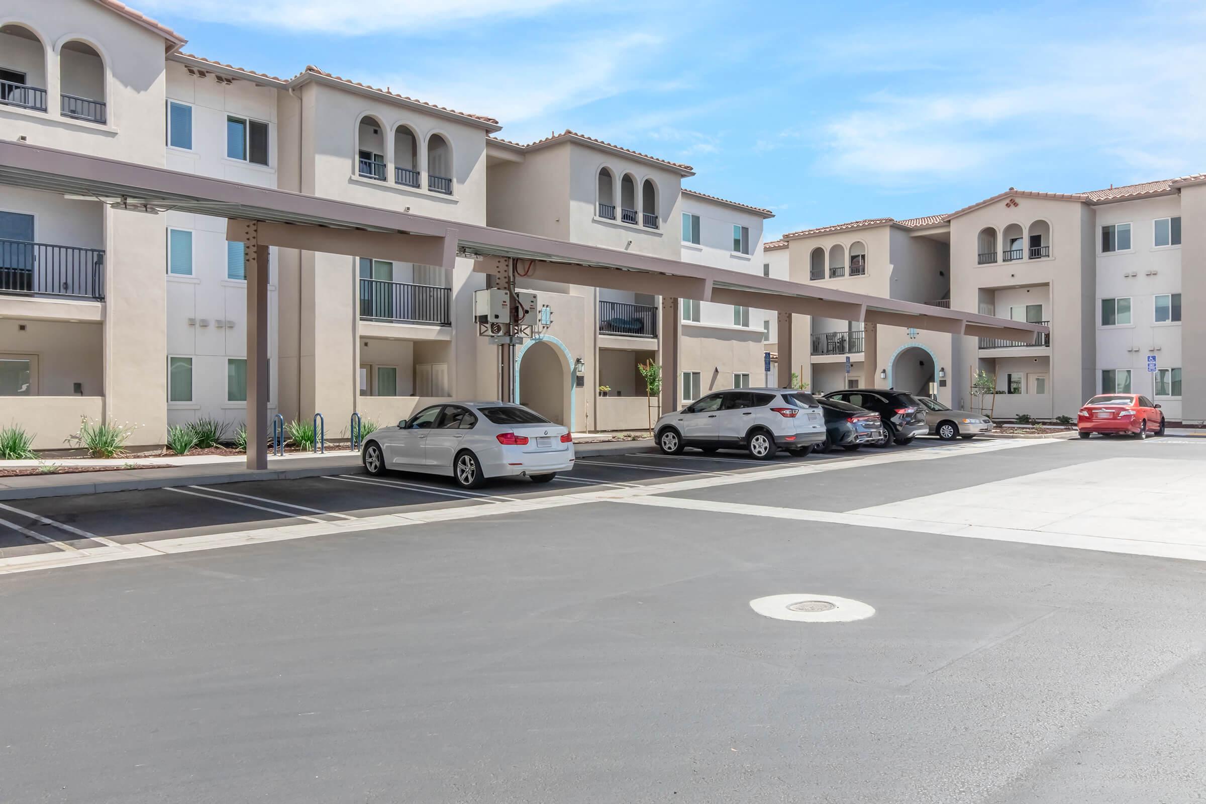 A residential apartment complex with multiple buildings featuring arches and balconies. The foreground shows a parking area with several parked cars under shaded structures. The background consists of landscaped areas, and the sky is clear and blue.