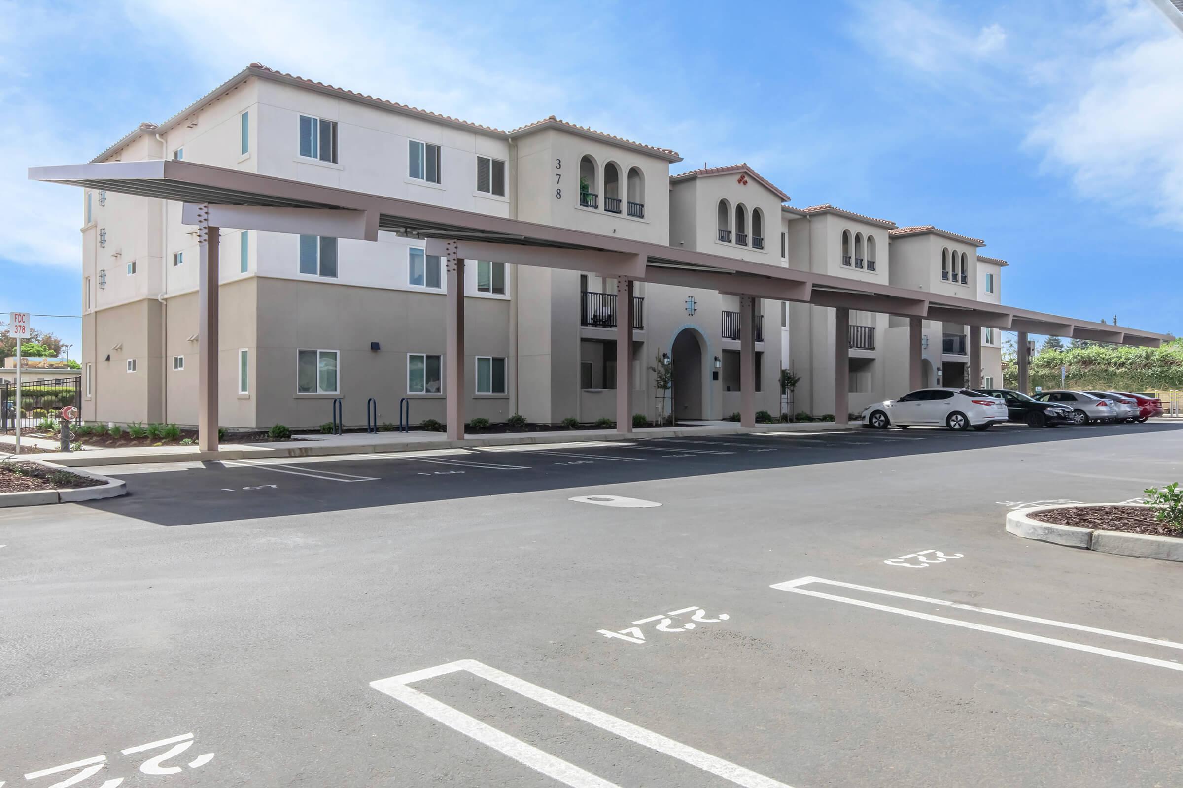 Three-story modern apartment building with a sandy-colored exterior and brown accents. There are covered parking spaces in the foreground, with several parked cars. The building features arched doorways and balconies, under a clear blue sky. Landscaping elements like small shrubs are visible along the perimeter.