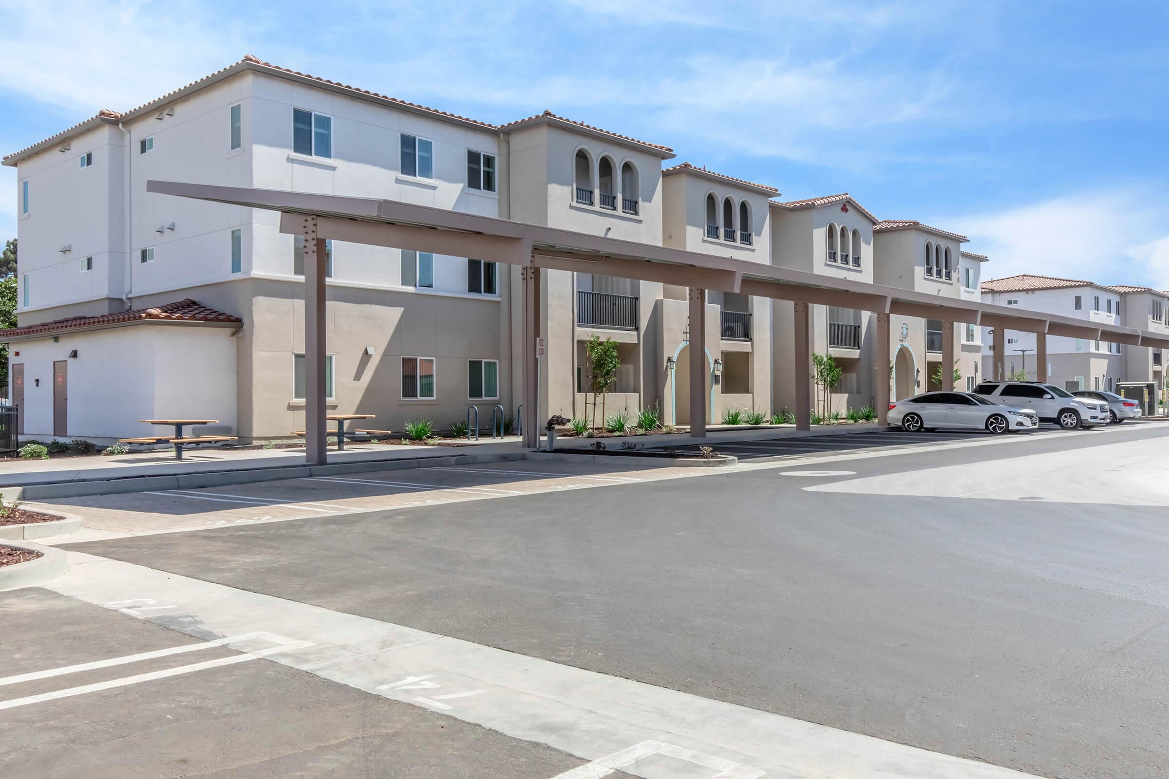 Modern apartment complex with beige and white exteriors, featuring multiple balconies and large windows. Covered parking spaces are visible in front, along with well-maintained landscaping. The sky is clear and blue, complementing the contemporary architecture.