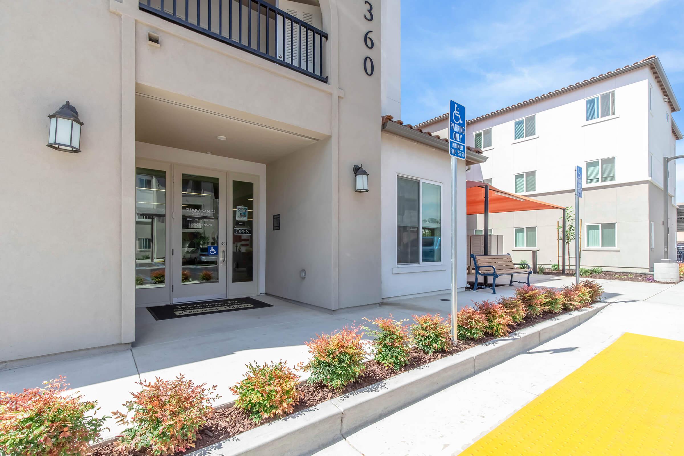 Entrance to a modern building with a welcoming porch area. The facade is light-colored with decorative light fixtures. Nearby, there are potted plants lining the walkway. A bench is visible, and a designated area for accessible parking is marked in the foreground. The sky is clear and blue, enhancing the overall inviting atmosphere.