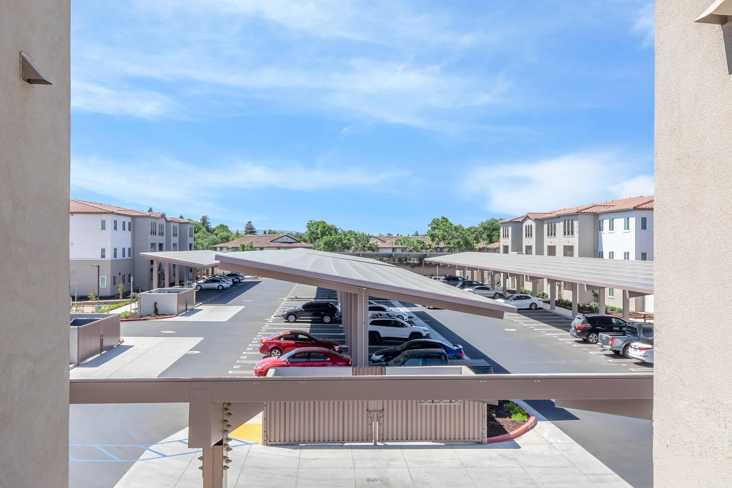 View from a balcony overlooking a parking lot filled with various cars, with modern apartment buildings in the background and clear blue skies. The scene captures a sunny day with well-maintained grounds and shaded parking areas.