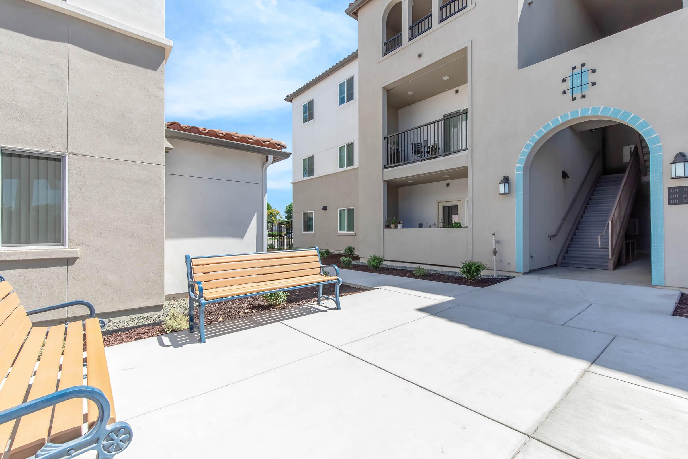Two benches are situated in a courtyard area, with a pathway leading to an entrance of a multi-story building. The building features a stairway, balconies, and light-colored exterior walls under a clear blue sky. Small shrubs are planted along the pathway.