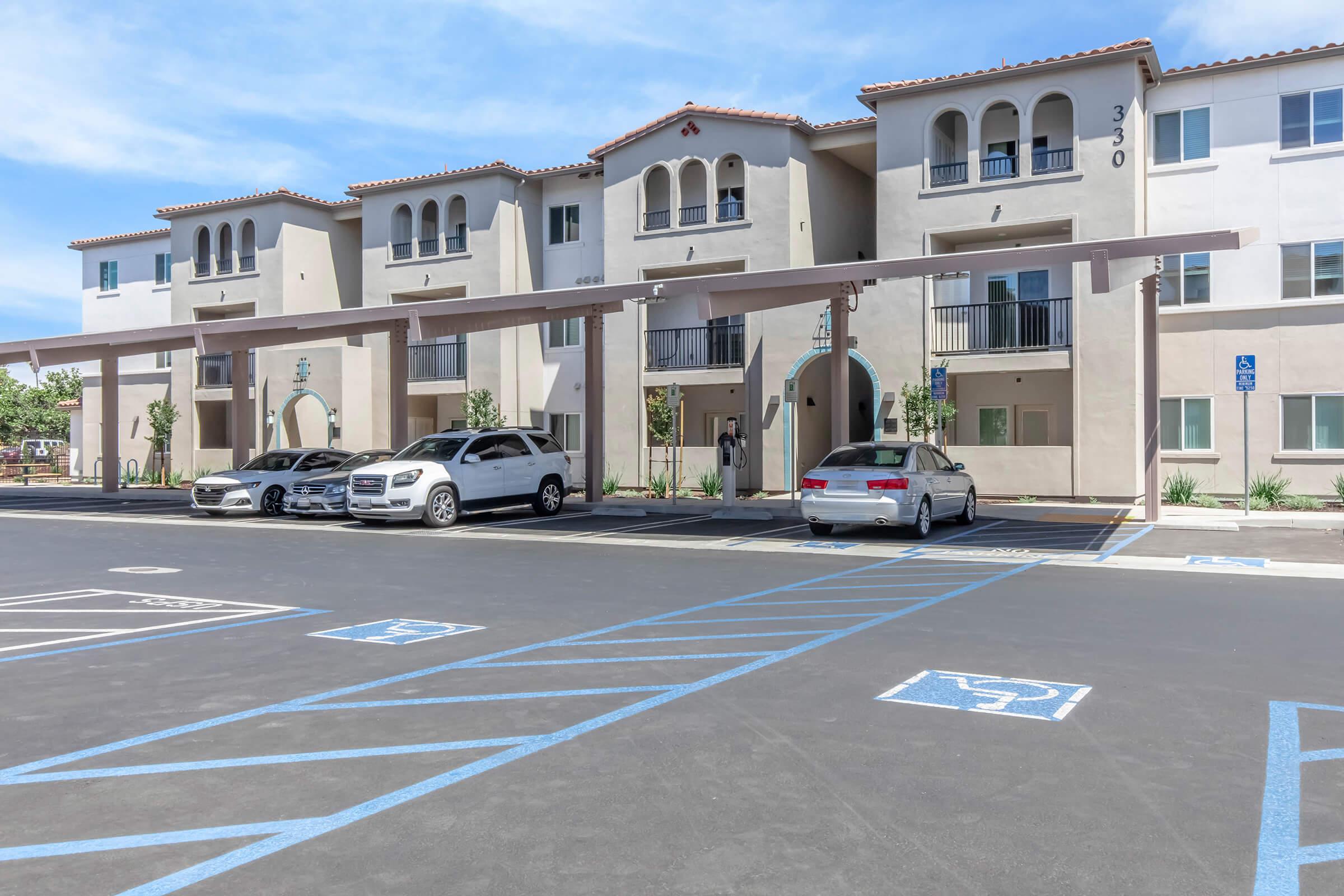 A modern residential building with a stucco exterior featuring arches and balcony spaces. Several vehicles are parked in front, including sedans and SUVs, under covered parking. The area is well-maintained with landscaping and clear blue skies in the background.