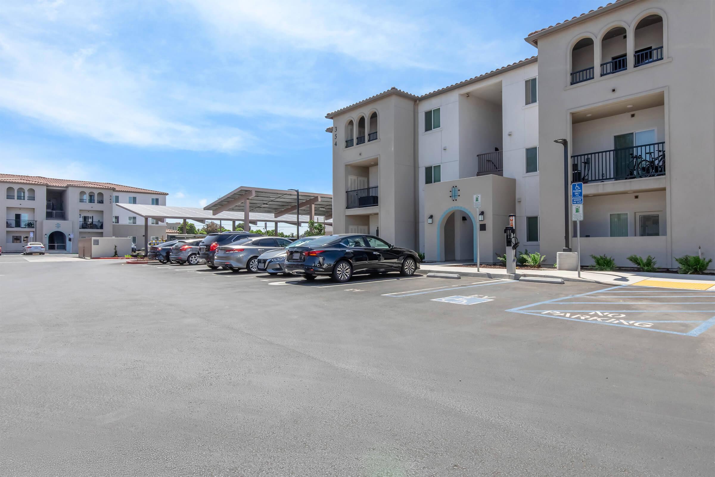 A modern apartment complex with three buildings featuring beige stucco exteriors. The parking lot is visibly empty with several parked cars. There are shaded areas for parking, and the sky is bright with a few clouds, indicating clear weather. Green landscaping is present near the buildings.