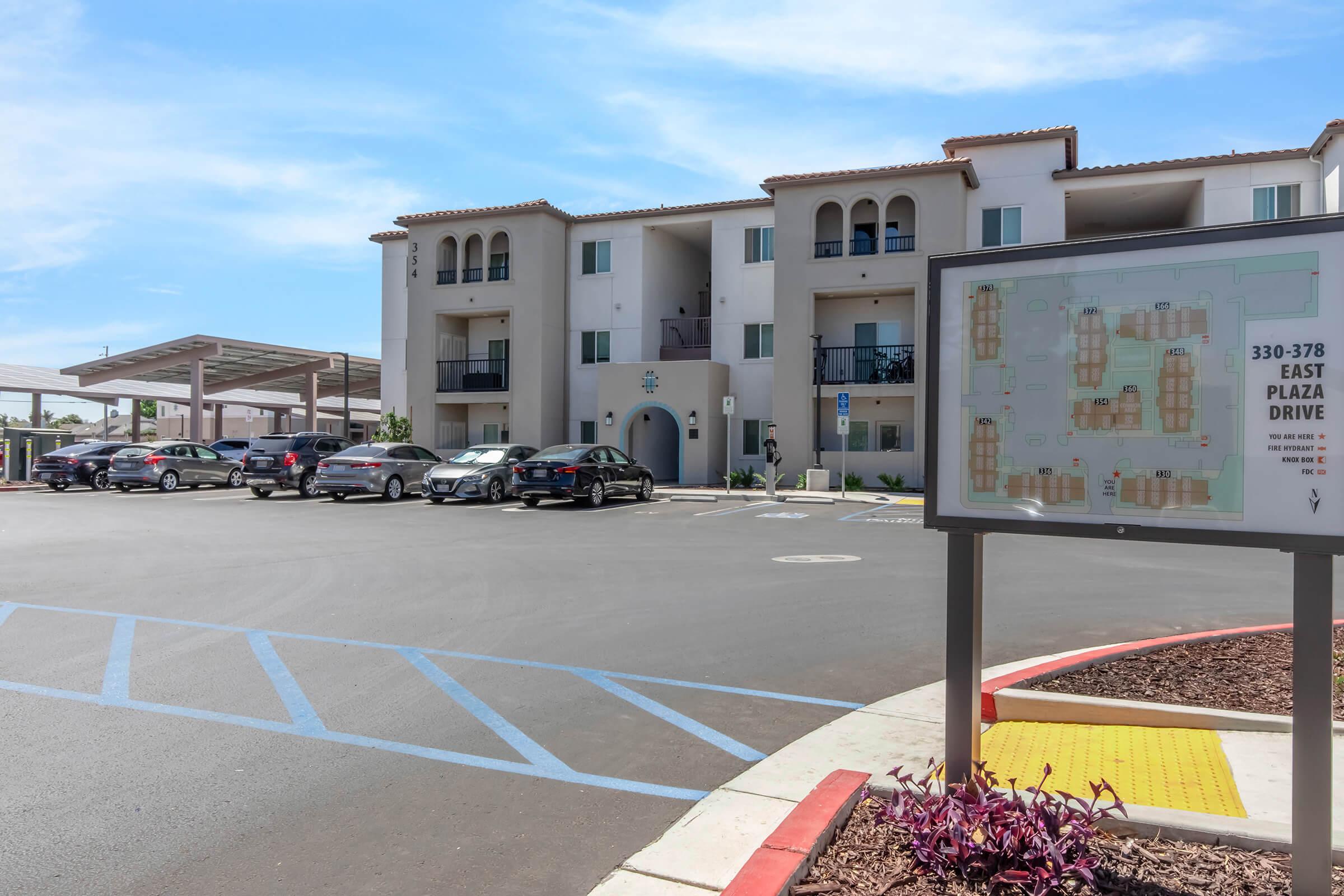 View of a modern apartment complex with a parking lot in the foreground. A large sign displays a map of the area, indicating various units and facilities. Several cars are parked in designated spaces, and there is ample blue sky overhead. The architecture features a Mediterranean style with arches and a stucco finish.