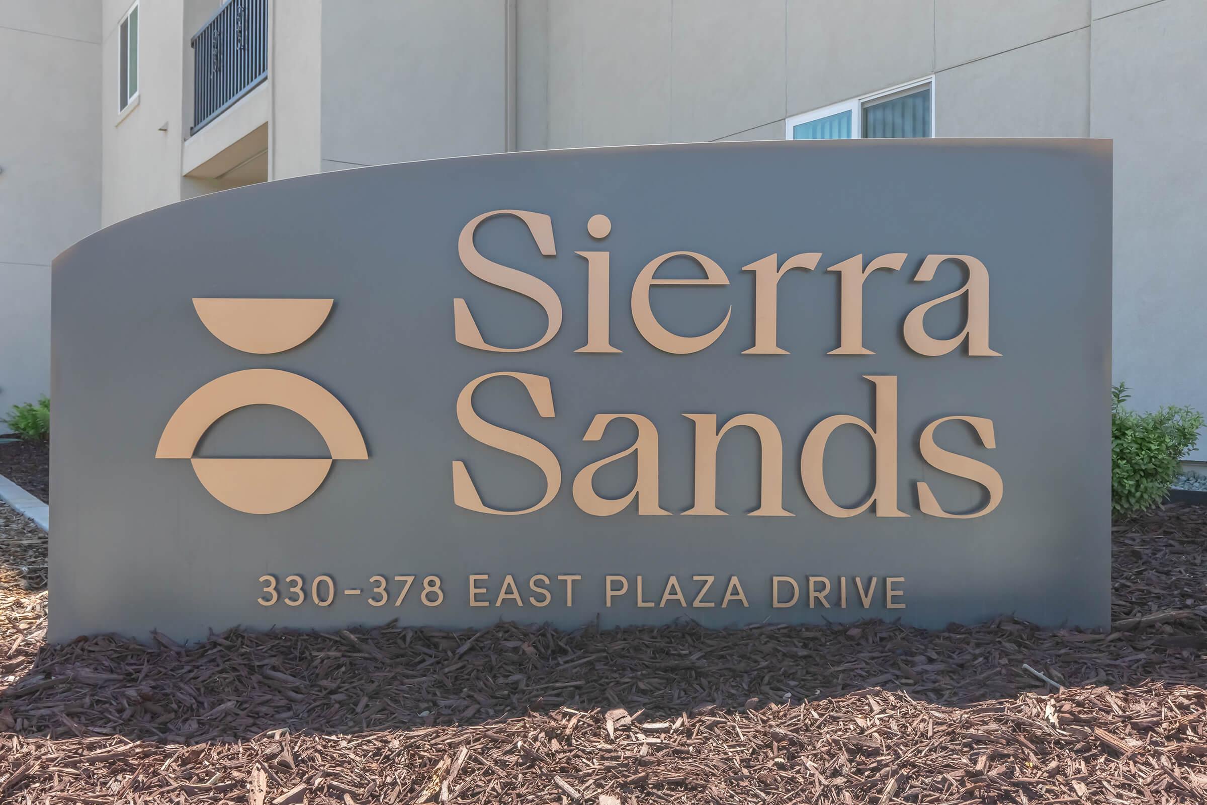Sign for Sierra Sands located at 330-378 East Plaza Drive. The design features a sleek, modern style with a two-part logo and the text prominently displayed in a warm color. Surrounding the sign is a landscaped area with mulch and some greenery.