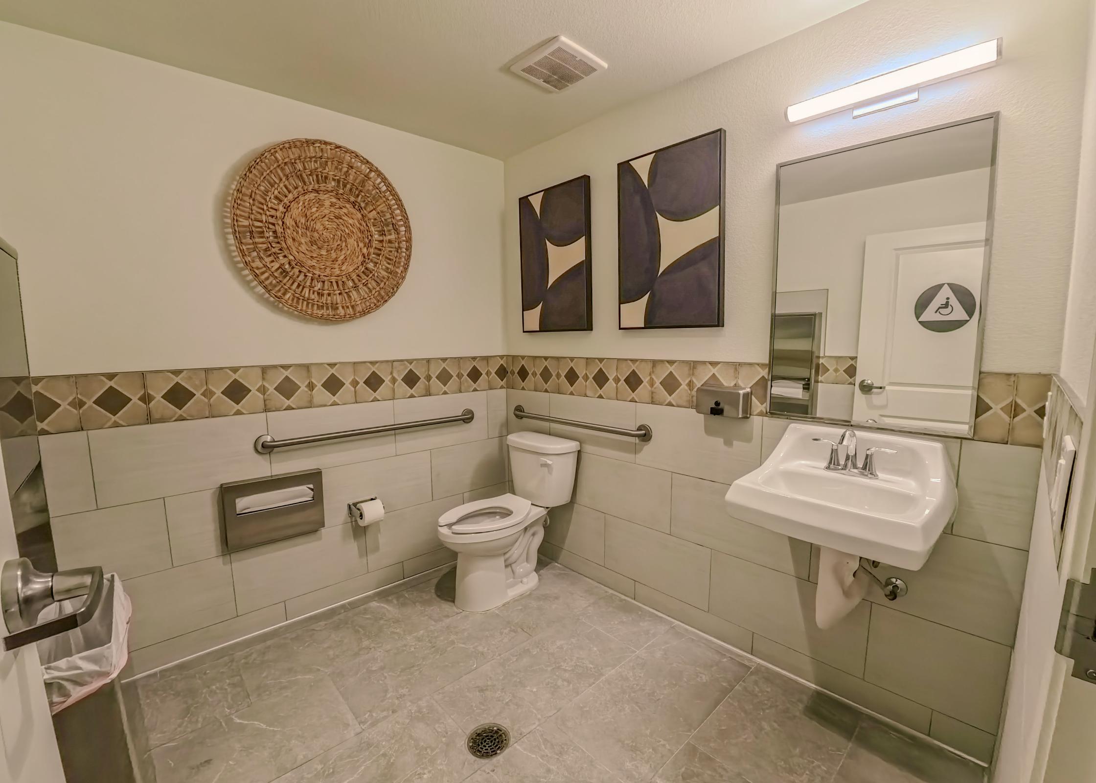 A clean, modern restroom featuring a toilet, sink, and wall-mounted mirror. The walls are adorned with minimalist artwork and decorative elements. Grab bars are installed for accessibility. The floor is tiled, and a trash bin is visible near the sink.