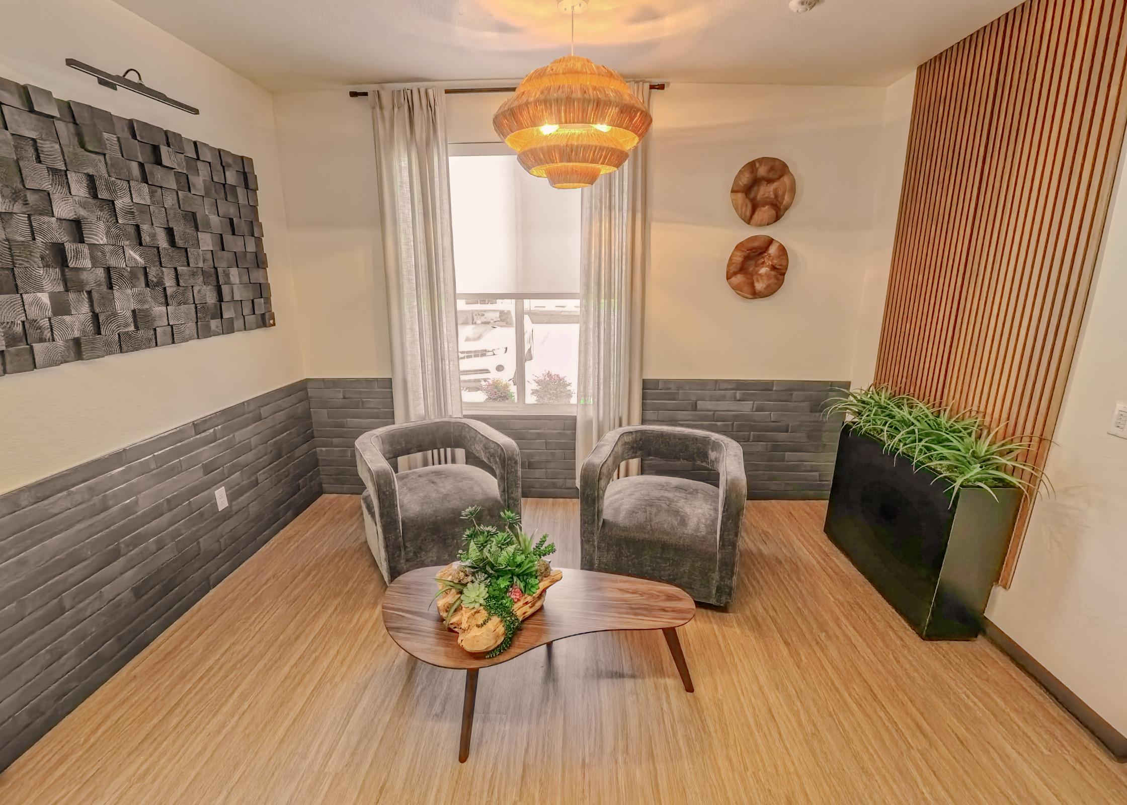 A cozy interior space featuring two gray armchairs facing a wooden coffee table with a plant center piece. Natural light filters through a window, with sheer curtains. The walls are partially decorated with textured panels, and there are decorative wall pieces and greenery adding warmth to the modern design.