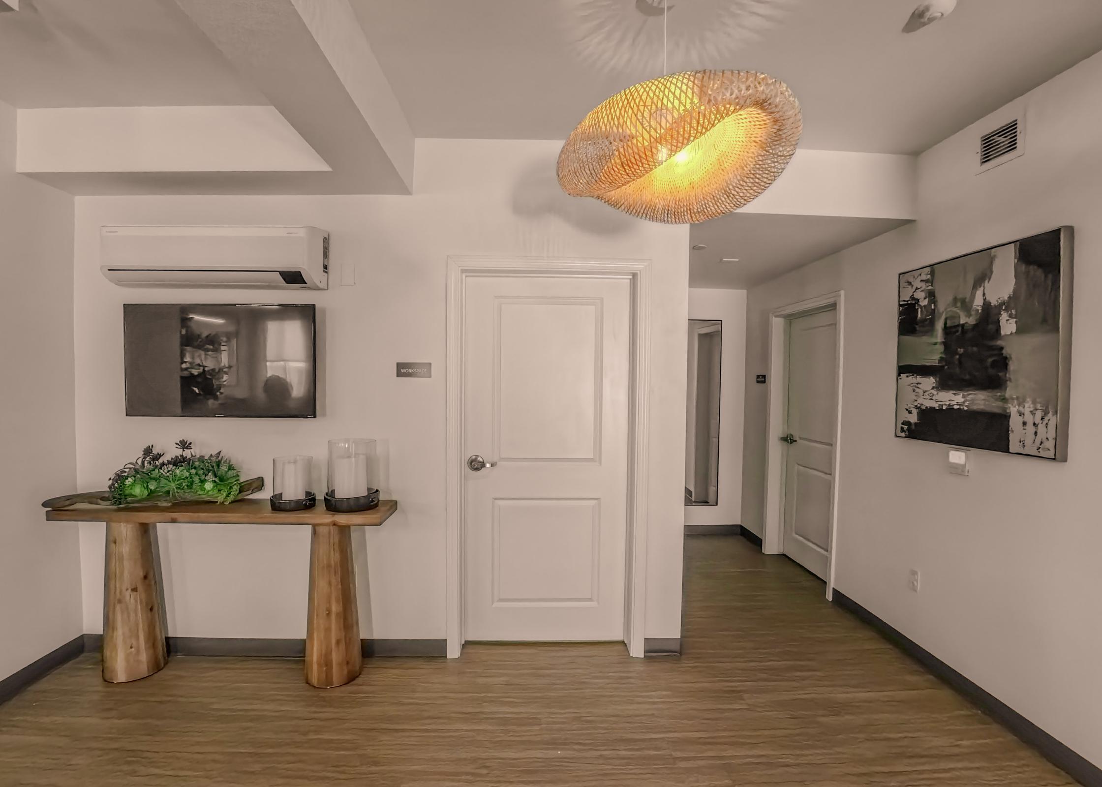 A modern interior space featuring a light wood table with decorative plants, a wall-mounted TV, and a stylish pendant light. The walls are painted white, and there are doorways leading to additional rooms. The flooring is light wood, creating a warm and inviting atmosphere.