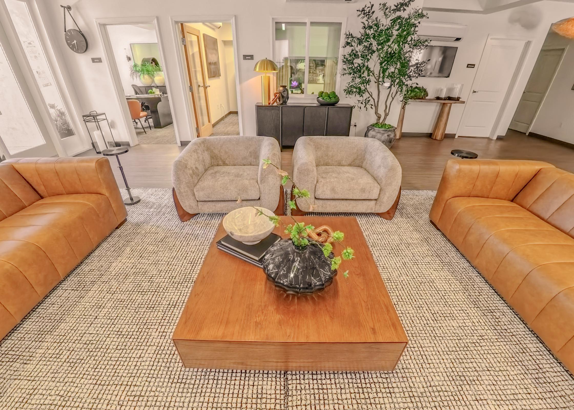 A cozy living room featuring two light brown armchairs and two tan sofas, arranged around a wooden coffee table. The table displays a decorative bowl and a plant. Surrounding the space are potted plants and a neutral-colored rug, with glimpses of doorways and a modern decor style in the background.
