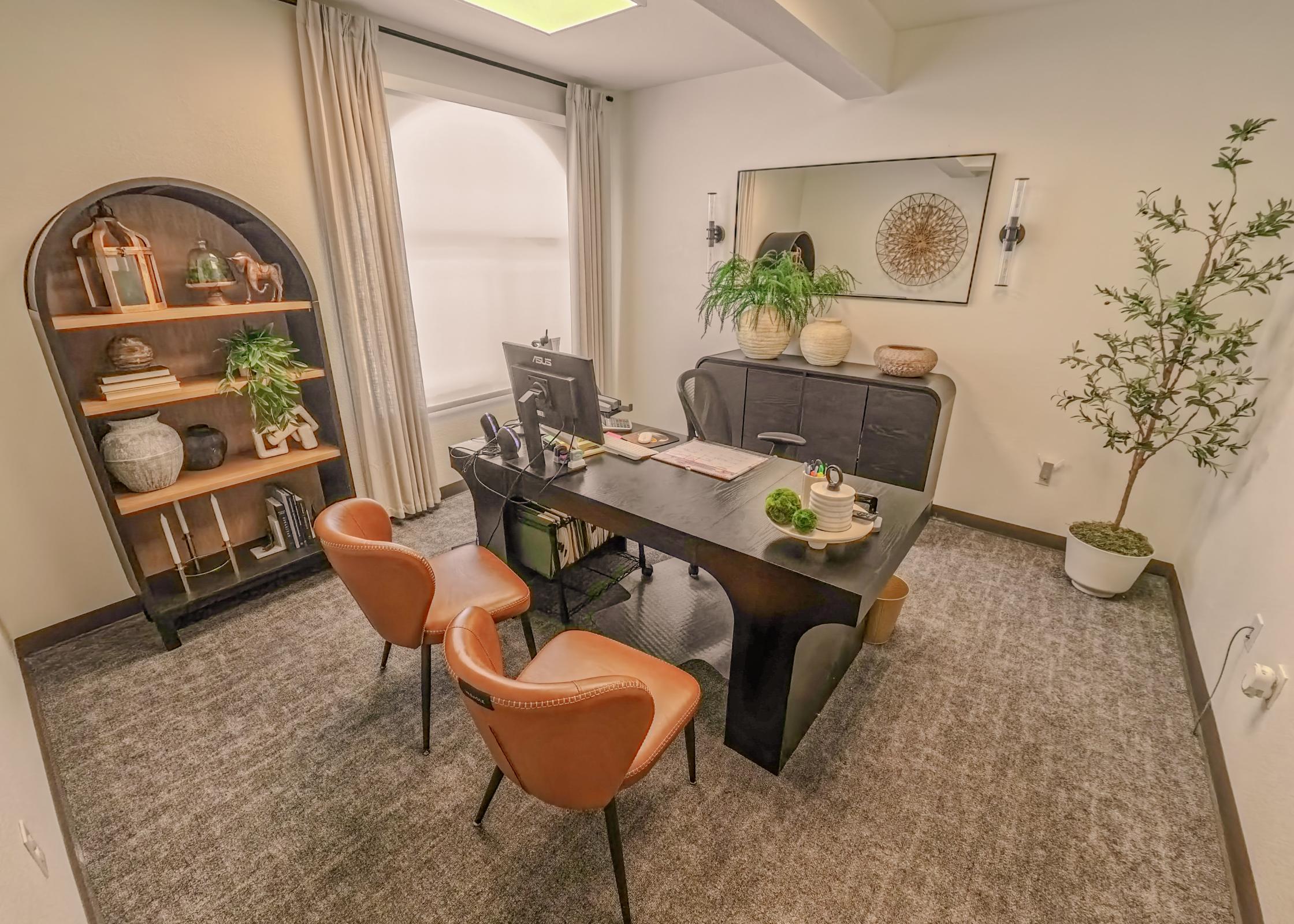 A stylish office space featuring a black desk with a computer, two brown leather chairs, and indoor plants. A wooden bookshelf displays decorative items and greenery. Large windows are dressed with light curtains, and a round mirror hangs on the wall, adding depth and warmth to the contemporary design.