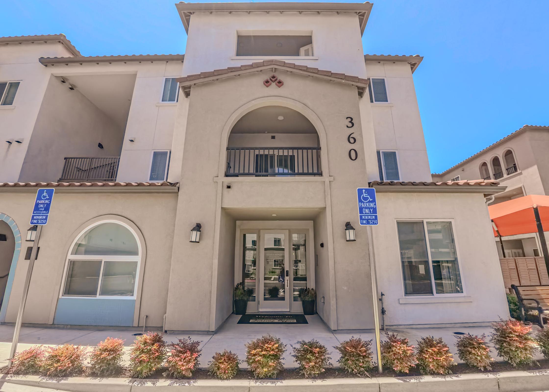 Exterior view of a modern apartment building featuring a prominent entrance with the number "360" displayed above. The façade is light-colored with a tile roof, and there are decorative plants in front. Accessibility signs are visible, and the setting is bright under a clear blue sky.