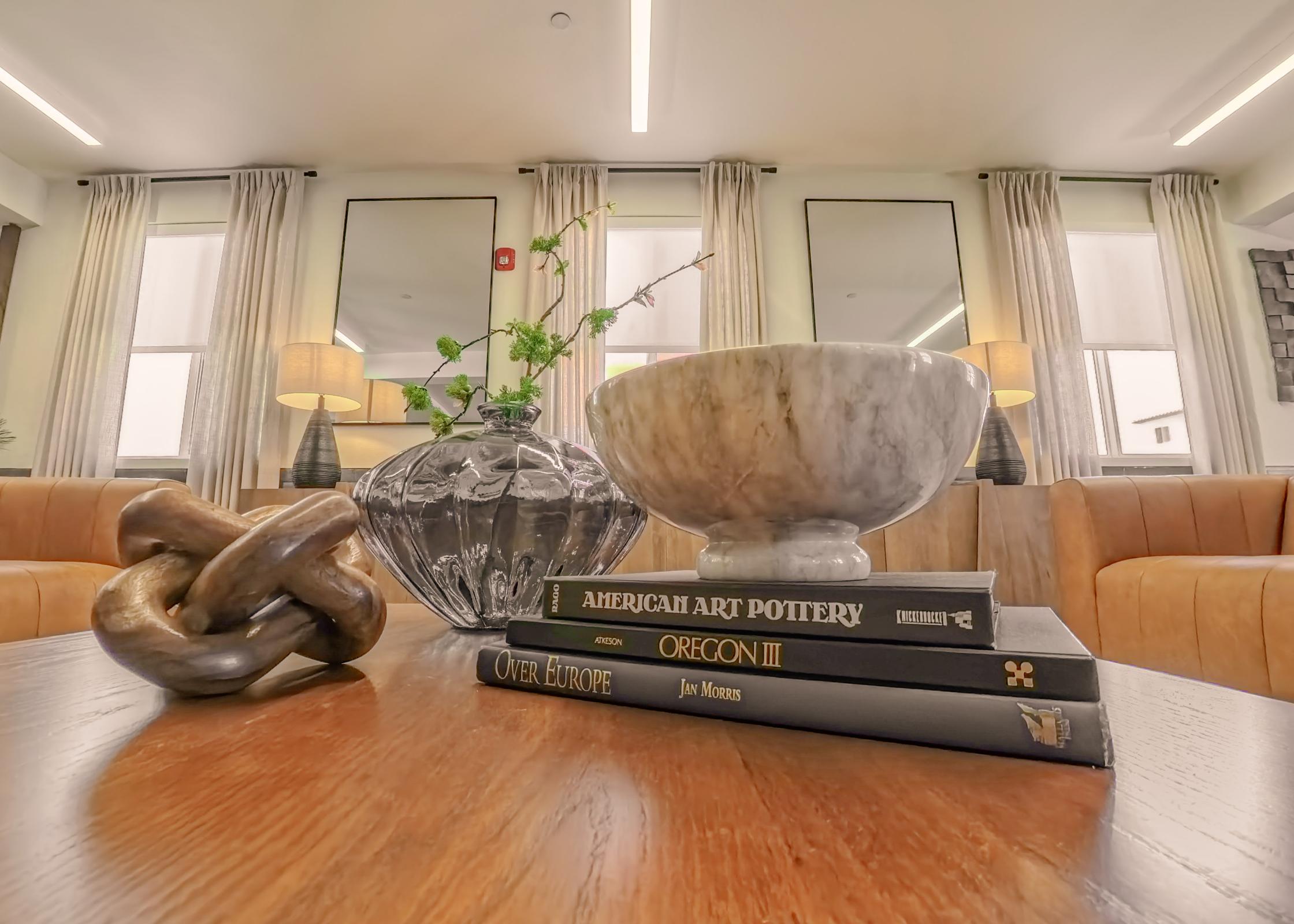 A stylish interior scene featuring a dark wooden table with stacked books titled "American Art Pottery" and "Oregon III." There's a decorative glass bowl, a small glass vase with greenery, and a wooden knot sculpture. The background showcases modern furniture, large windows, and stylish lamps, creating a cozy atmosphere.