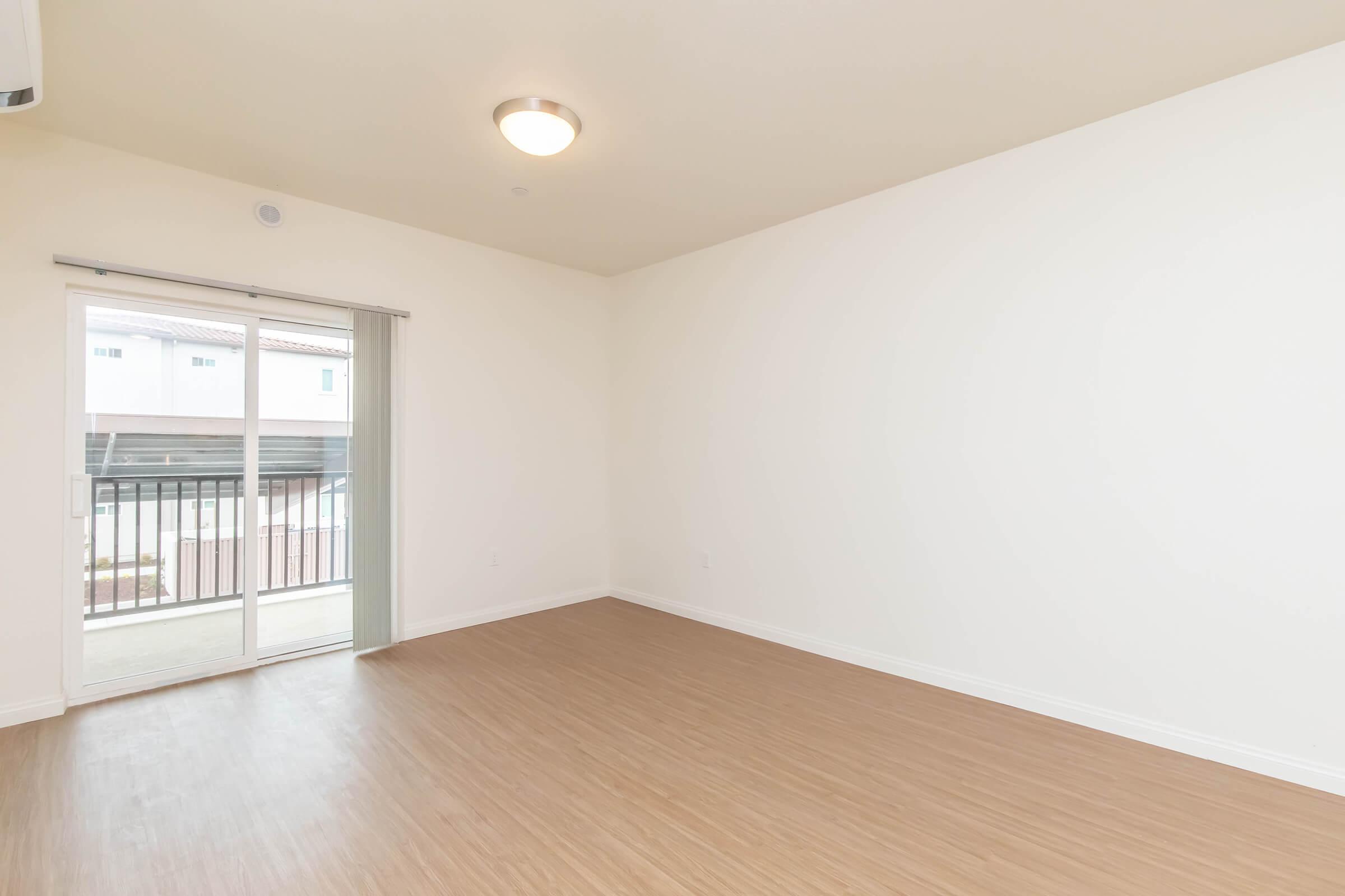 A spacious, empty room with light-colored walls and a wooden floor. A sliding glass door leads to a small balcony, letting in natural light. The room is unadorned, presenting a clean, minimalist aesthetic, ideal for personalization.