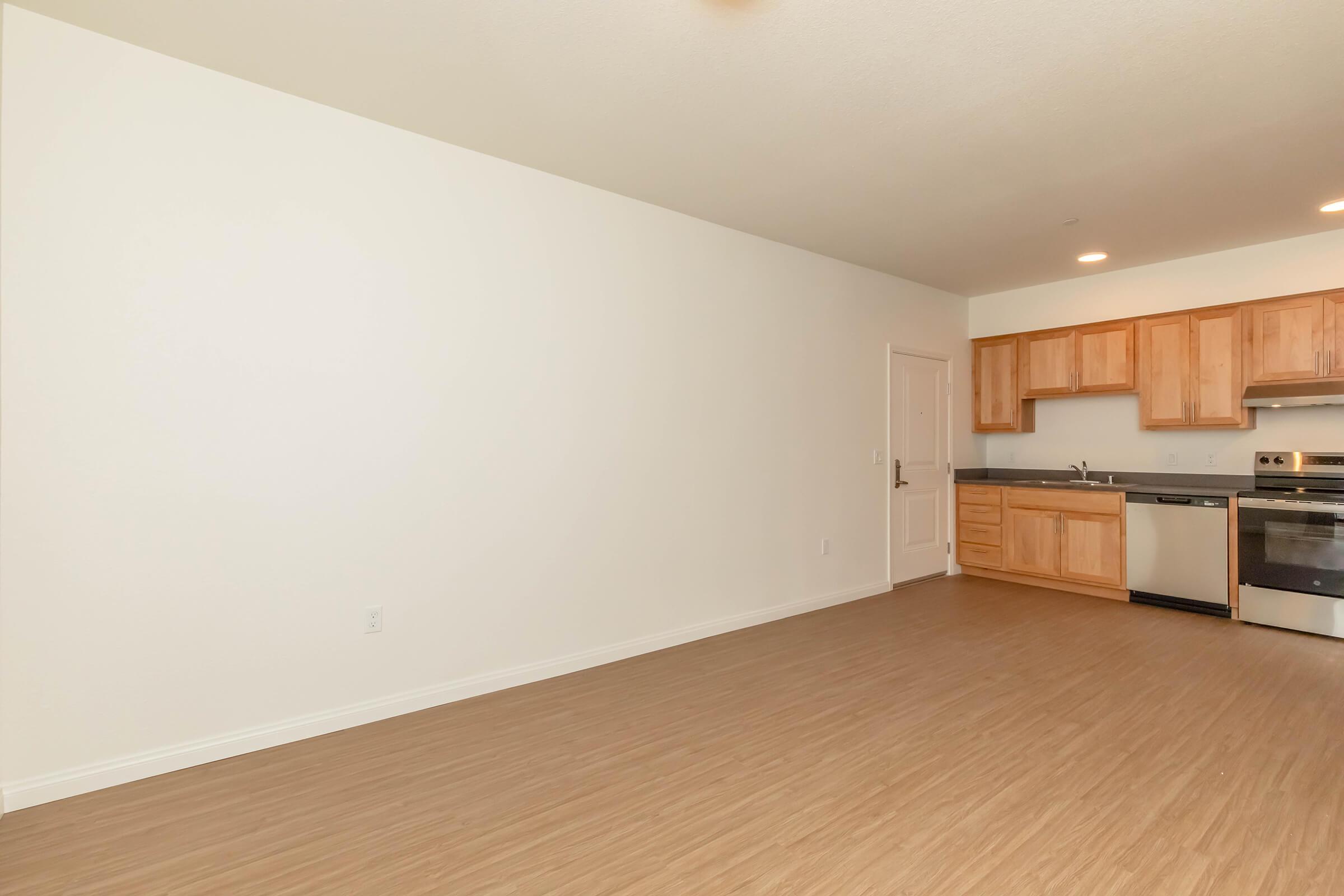 An empty room showcasing a minimalist design with light wooden flooring. On one side, there is a kitchen area featuring wooden cabinets, stainless steel appliances, and a countertop. The walls are painted white, creating a bright and airy atmosphere. The space appears clean and well-lit.