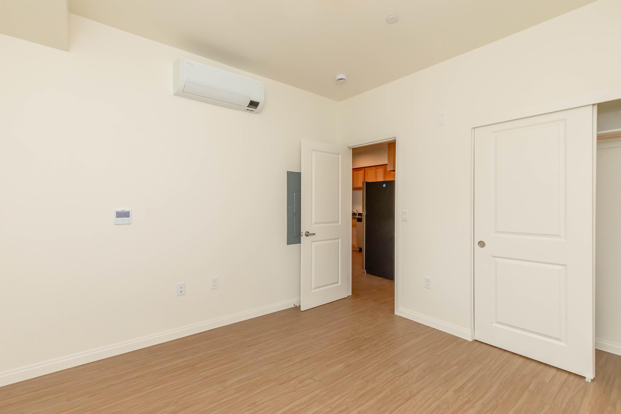 A small, empty room with light-colored walls and wooden flooring. It features a wall-mounted air conditioning unit, an open door leading to a kitchen area, and a closet with a closed door. The space is well-lit and has a minimalist design.