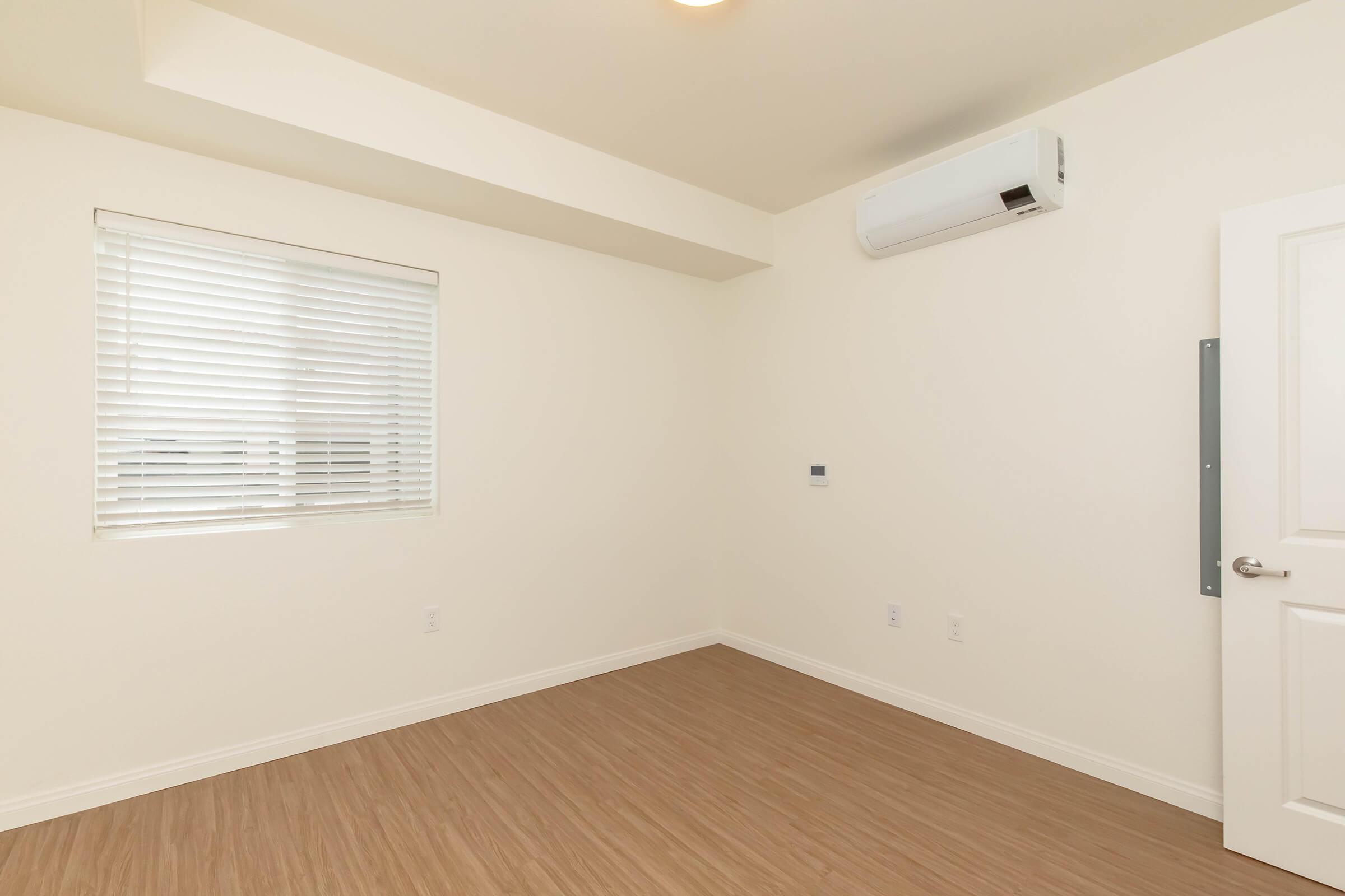 A bright, empty room featuring light-colored walls, a window with white blinds, and a wood-style floor. An air conditioning unit is mounted on the wall, and a white door is partially visible on the right side. The overall space is minimalist and modern.