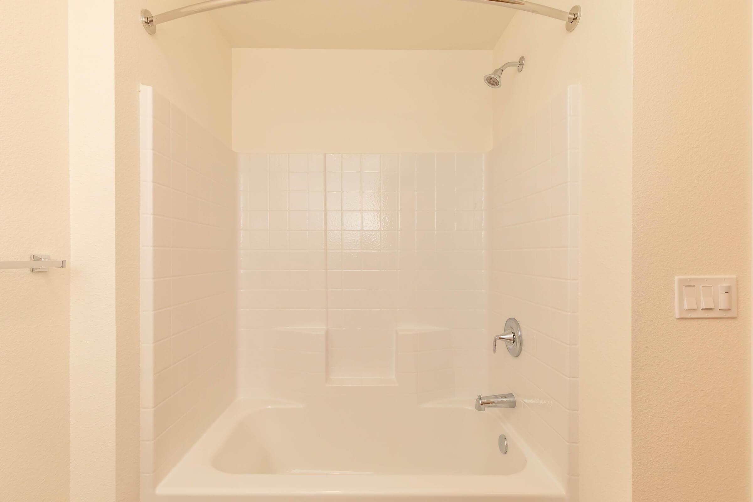 A clean, empty bathtub with white tiled walls and a silver showerhead. The bathroom features a light neutral-colored wall and a chrome towel bar. A single light switch is visible on the right side. The overall appearance is simple and modern.