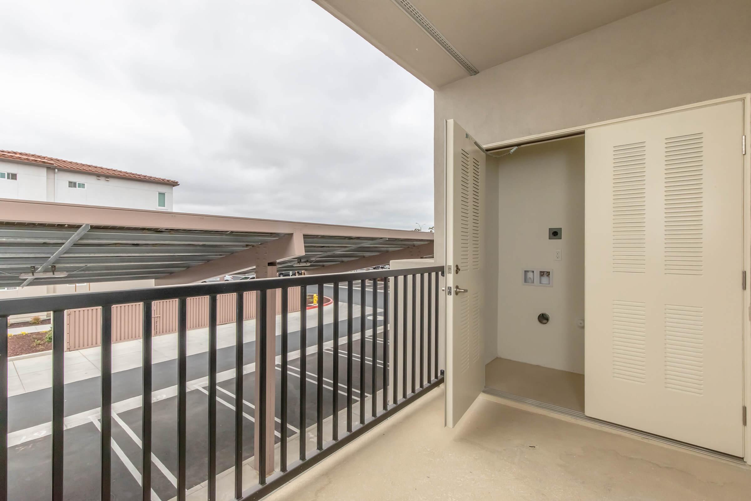 An open door on a balcony, revealing an interior space. The balcony has a railing and overlooks a parking area with shaded spots. The sky is overcast with clouds, and nearby buildings are visible in the background.