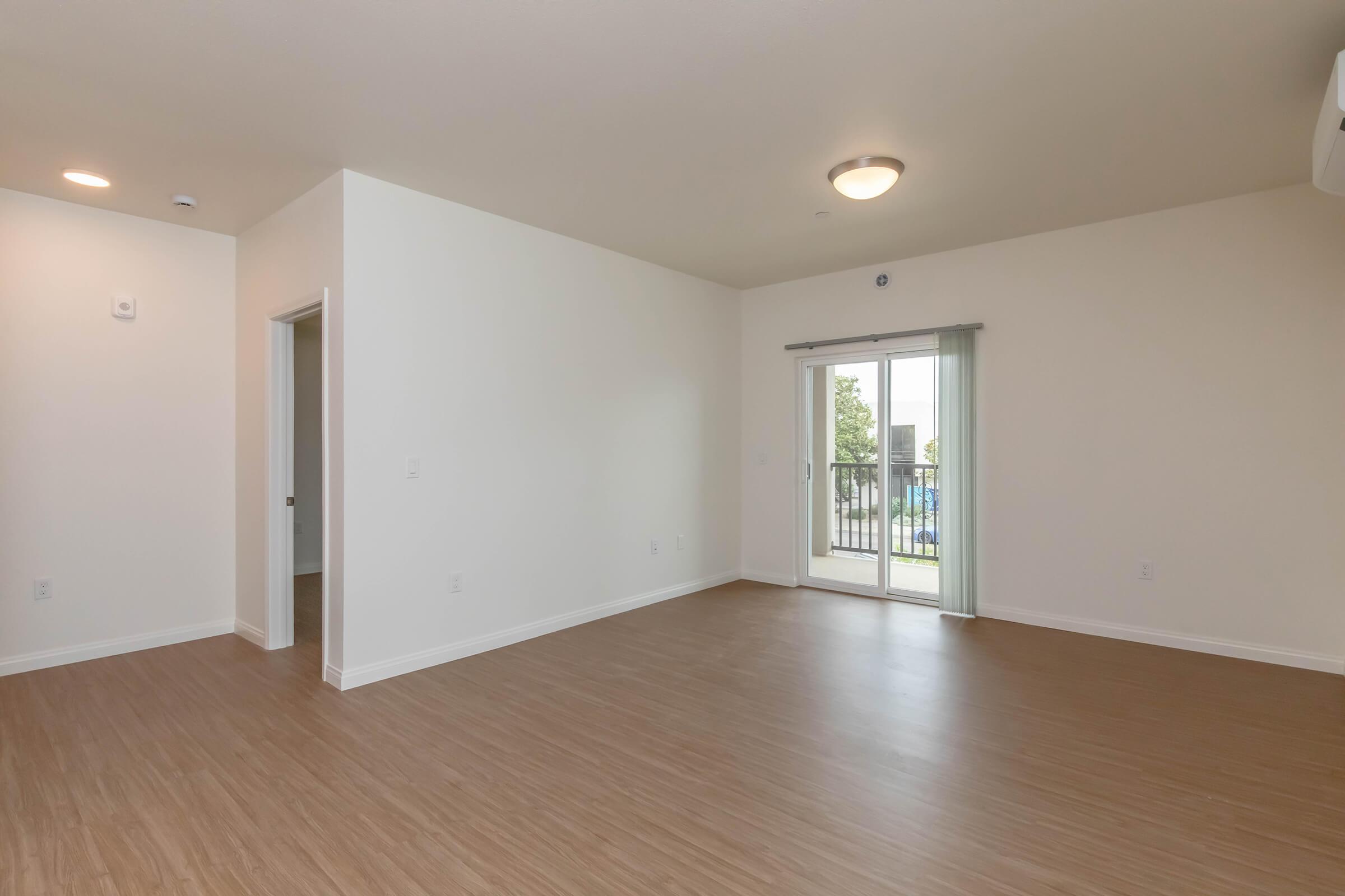 A spacious, empty room with light-colored walls and wooden flooring. There is a sliding glass door leading to a balcony, and a small window. The room is well-lit, featuring a ceiling light fixture. The doorway on the left side leads to another room.