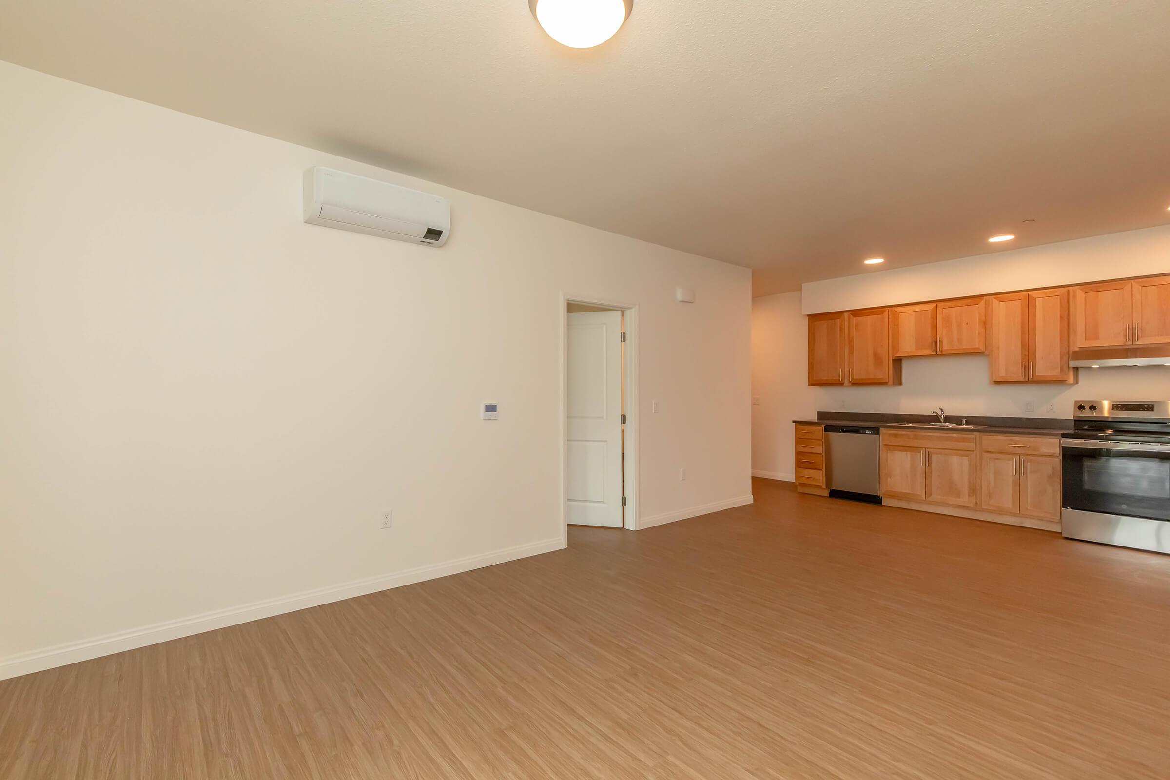 Spacious light-filled room featuring beige walls and wooden flooring, with an air conditioning unit on the wall. A doorway leads to another area. The kitchen is visible with wooden cabinets, stainless steel appliances, and a countertop, creating an open-concept living space.