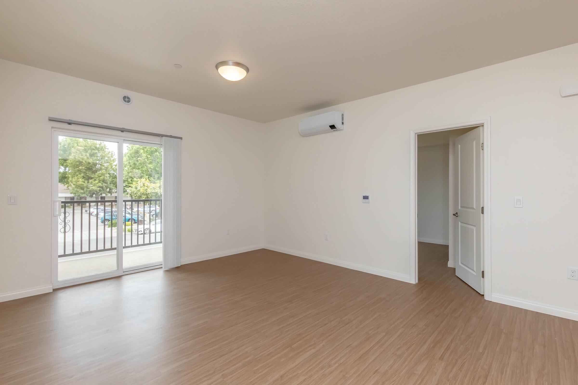A spacious, empty room with light-colored walls and a wood-like laminate floor. There is a sliding glass door leading to a small balcony, and a white air conditioning unit mounted on the wall. A door on the right leads to another room or area. Natural light floods the space from outside.