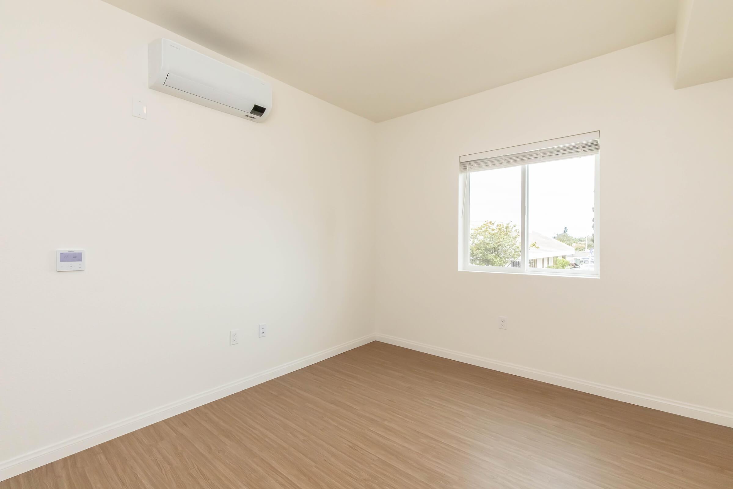 Empty room with light-colored walls and a window. The floor is made of light wood or laminate, and there is a wall-mounted air conditioning unit. A small control panel is visible on one wall. Natural light enters through the window, highlighting the spaciousness of the room.