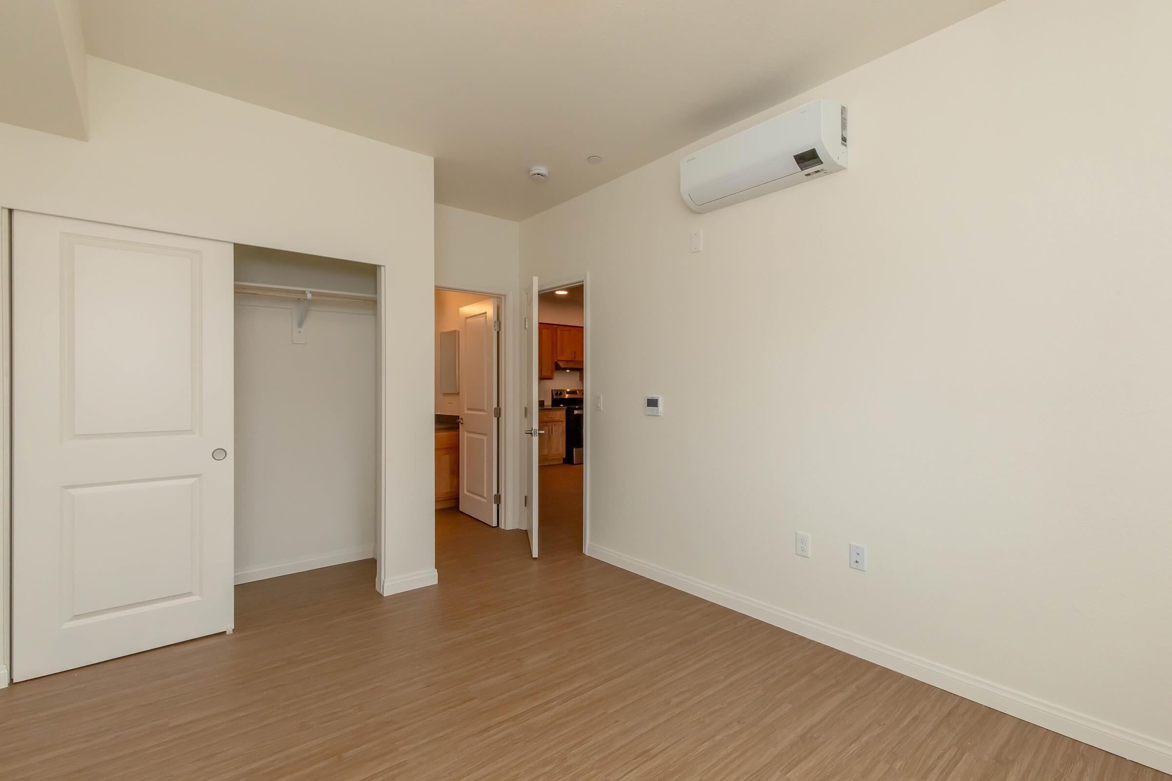 Interior view of a well-lit, empty room featuring light-colored walls and wooden flooring. There is a closed door leading to a kitchen area, a closet with sliding doors on one side, and a wall-mounted air conditioning unit. The space appears new and minimally decorated, suitable for personal furnishings.
