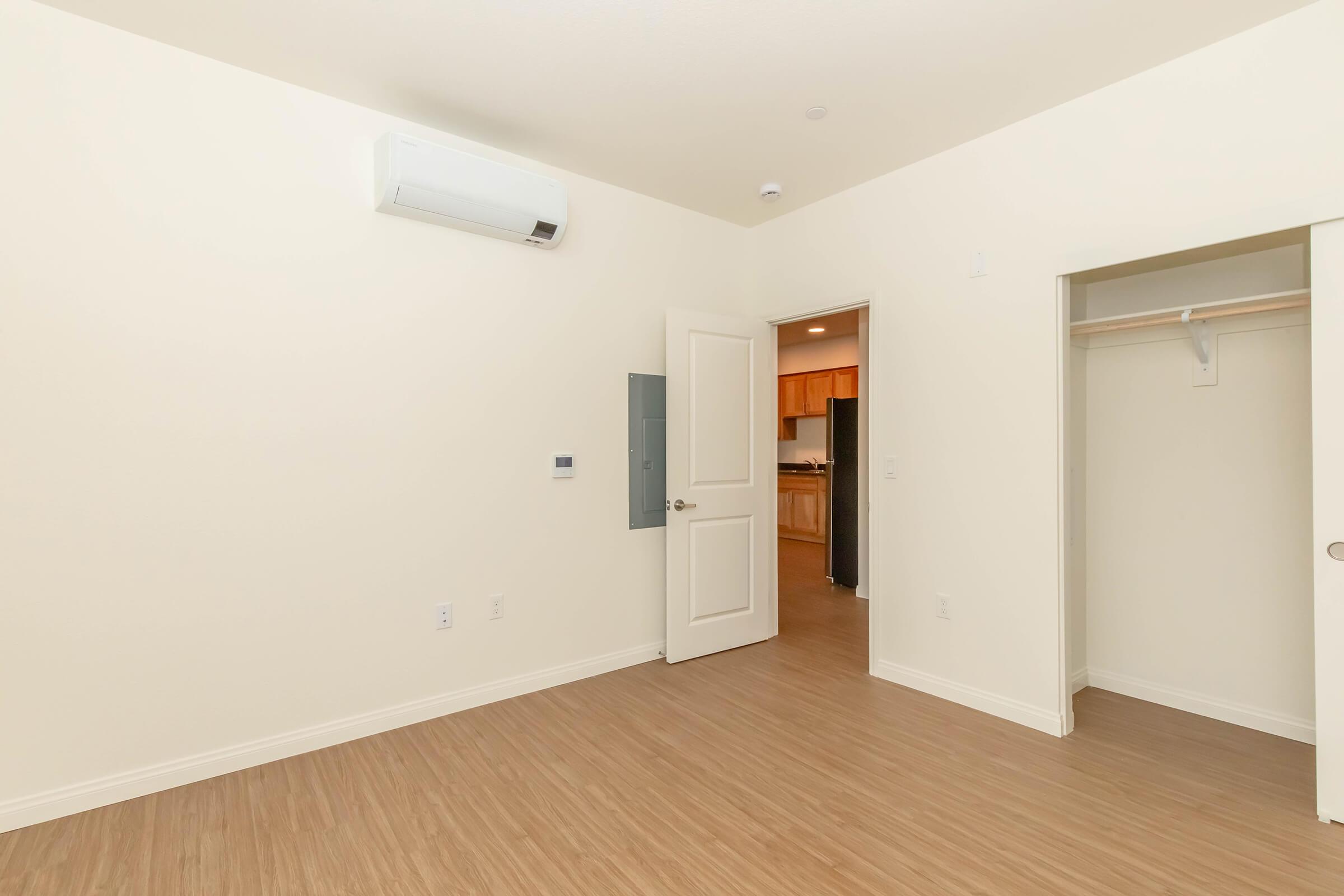 A vacant room with light-colored walls and wooden flooring. An air conditioning unit is mounted on one wall. A door leads to a kitchen area in the background, and there is a closet with sliding doors on the left. The space is well-lit and uncluttered, offering a minimalist aesthetic.