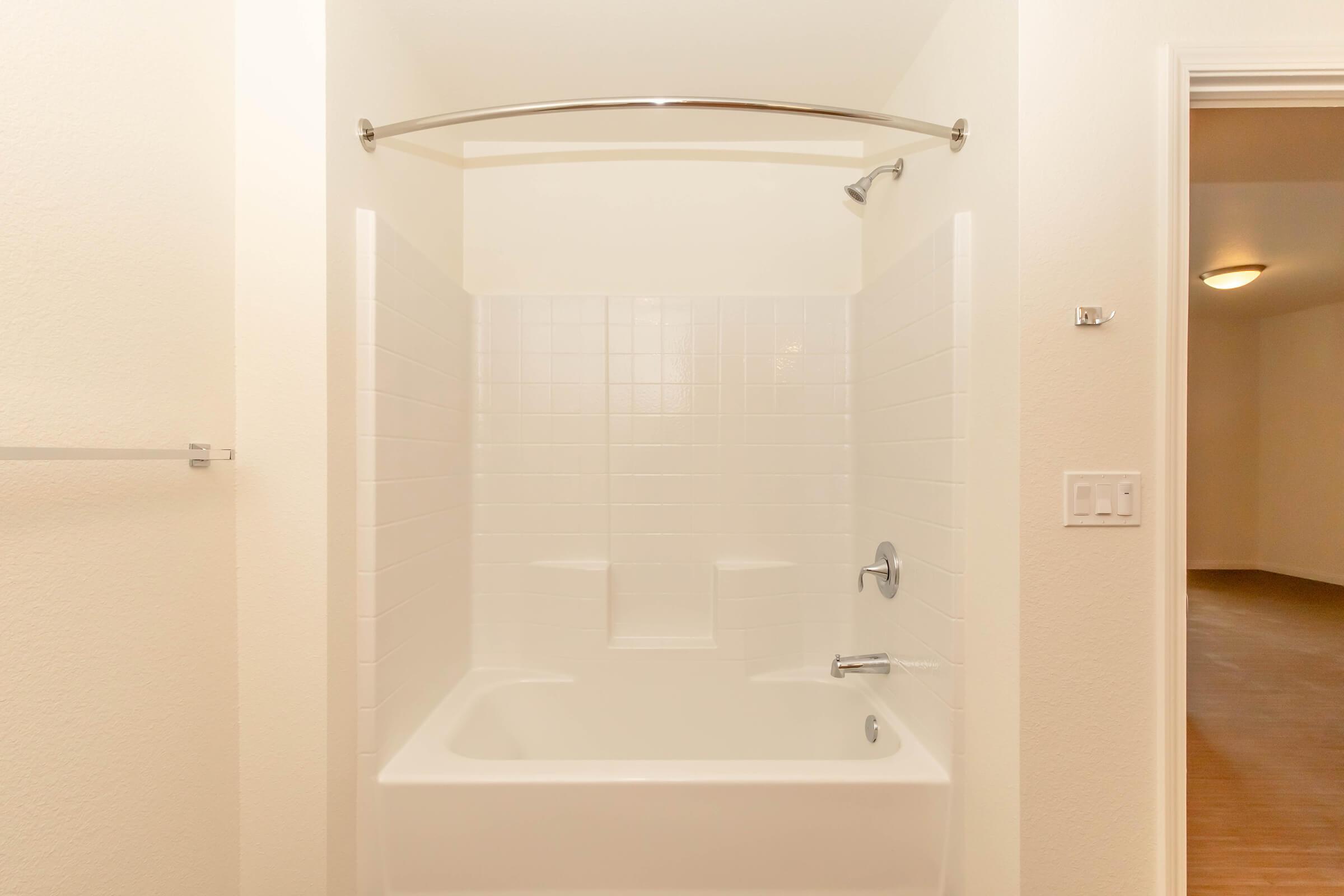 A clean, white bathroom featuring a bathtub with a curved shower rod. The walls are tiled with white ceramic tiles, and there are faucets and a showerhead visible. The doorway to another room is partially visible on the right, and the floor has a warm wooden finish.