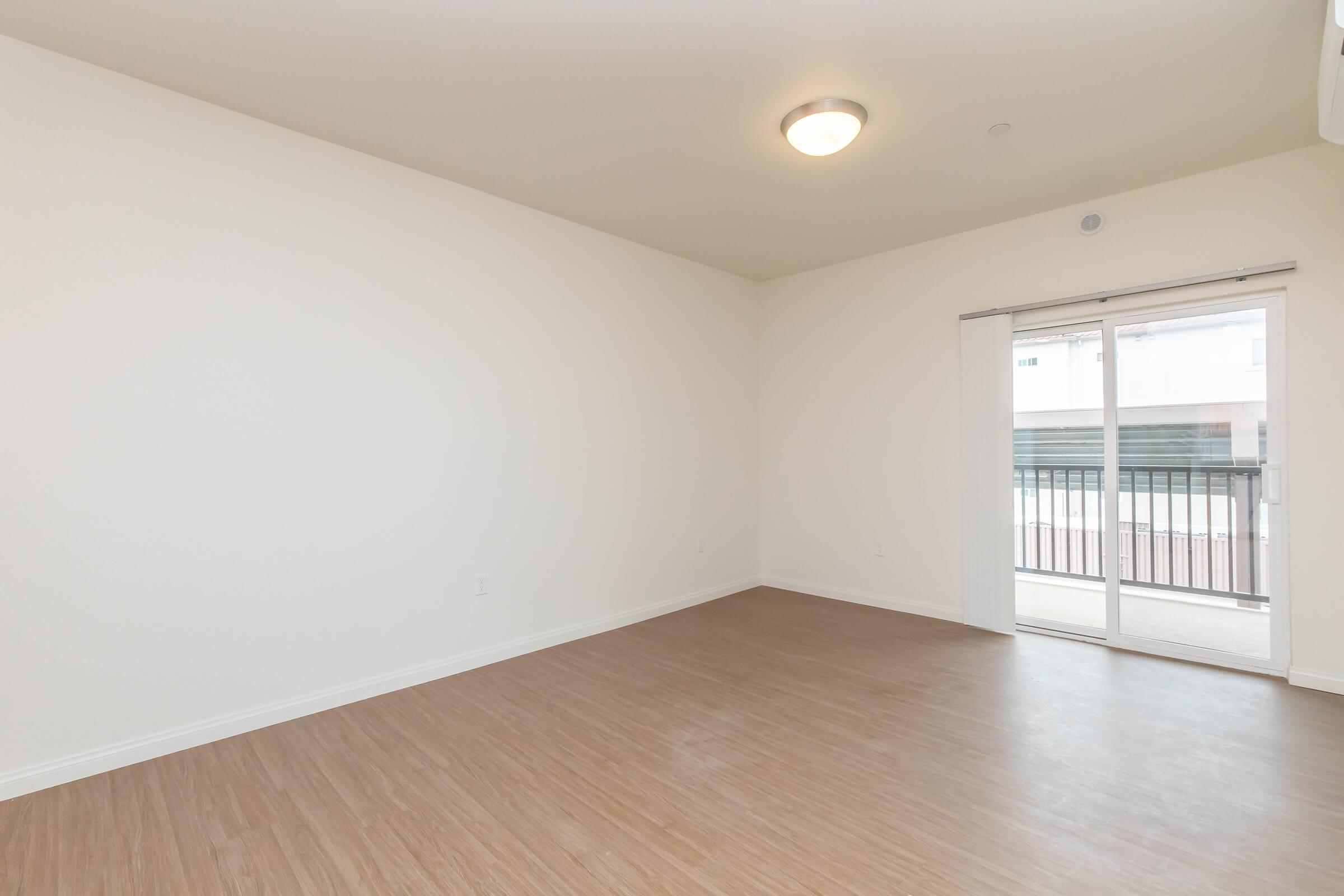 Spacious, empty room with light-colored walls and flooring. A sliding glass door leads to a balcony, allowing natural light into the space. The ceiling features a simple, modern light fixture. Ideal for various layout possibilities in a home or apartment setting.