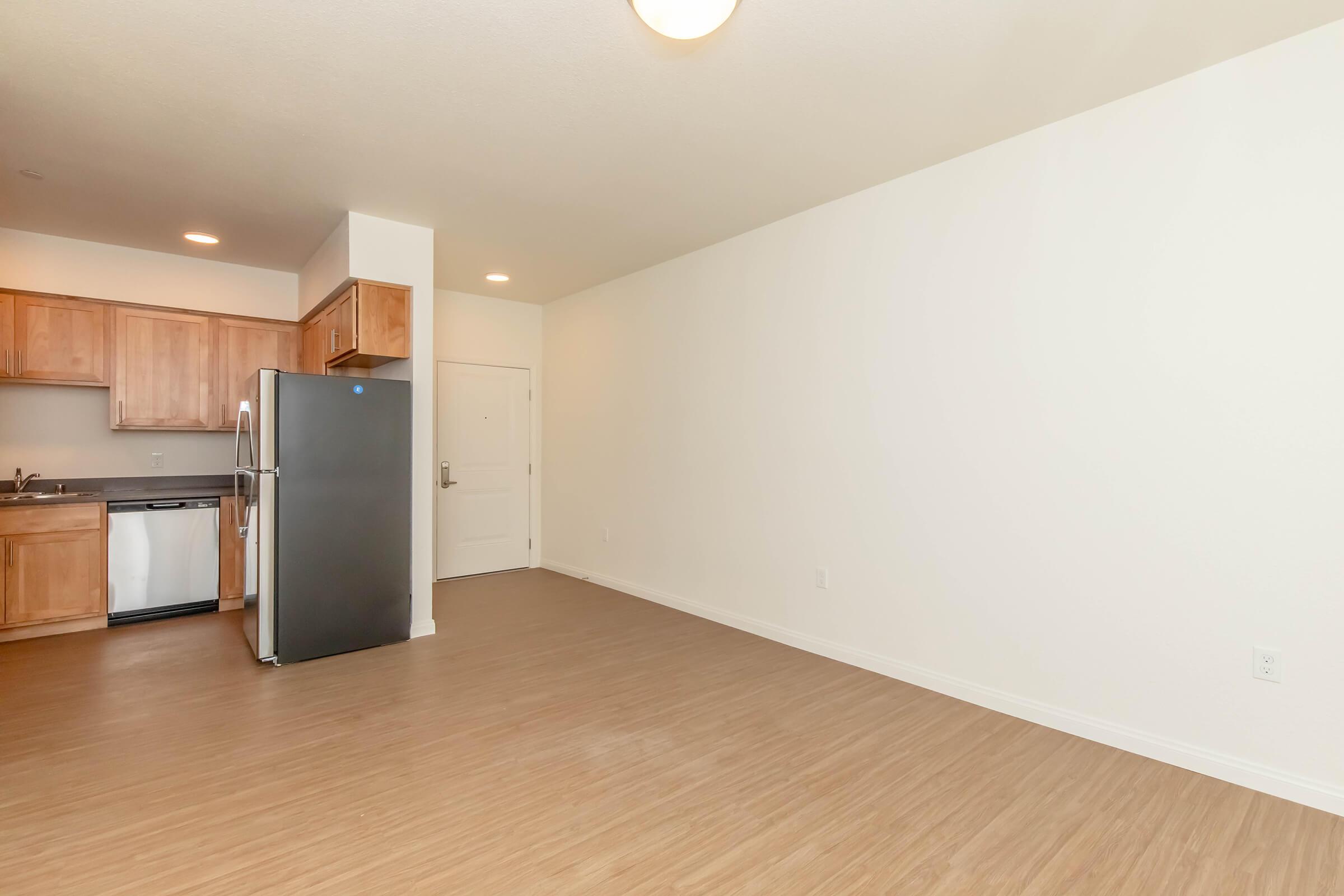 Interior of an empty apartment showing a kitchen area with wooden cabinets and a black refrigerator. A spacious living area is visible with light-colored walls and a smooth floor. The entry door is also seen, providing a bright and modern ambiance.