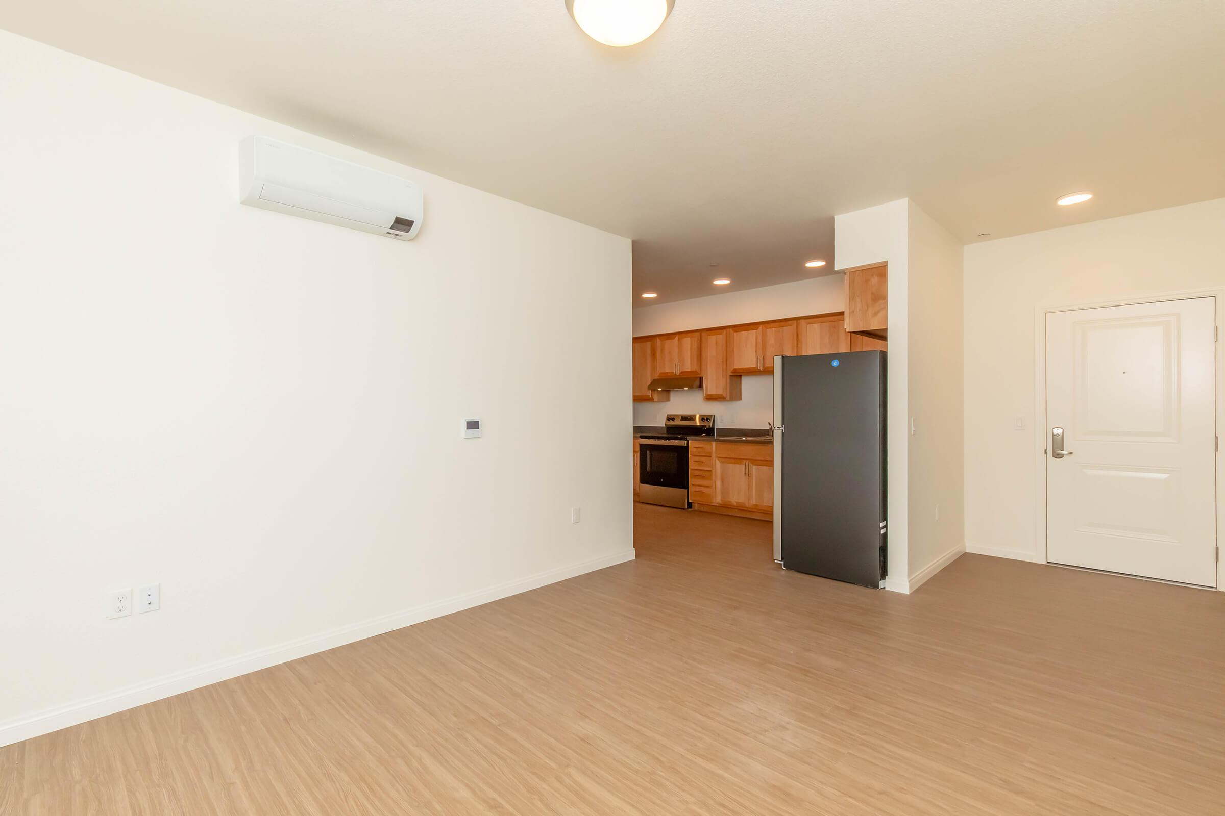 An empty living space featuring light-colored walls and wood-style flooring. To the right, there’s a kitchen area with wooden cabinets and stainless steel appliances, including a fridge. An entrance door is visible on the right. An air conditioning unit is mounted on the wall. The room is well-lit with ceiling lights.
