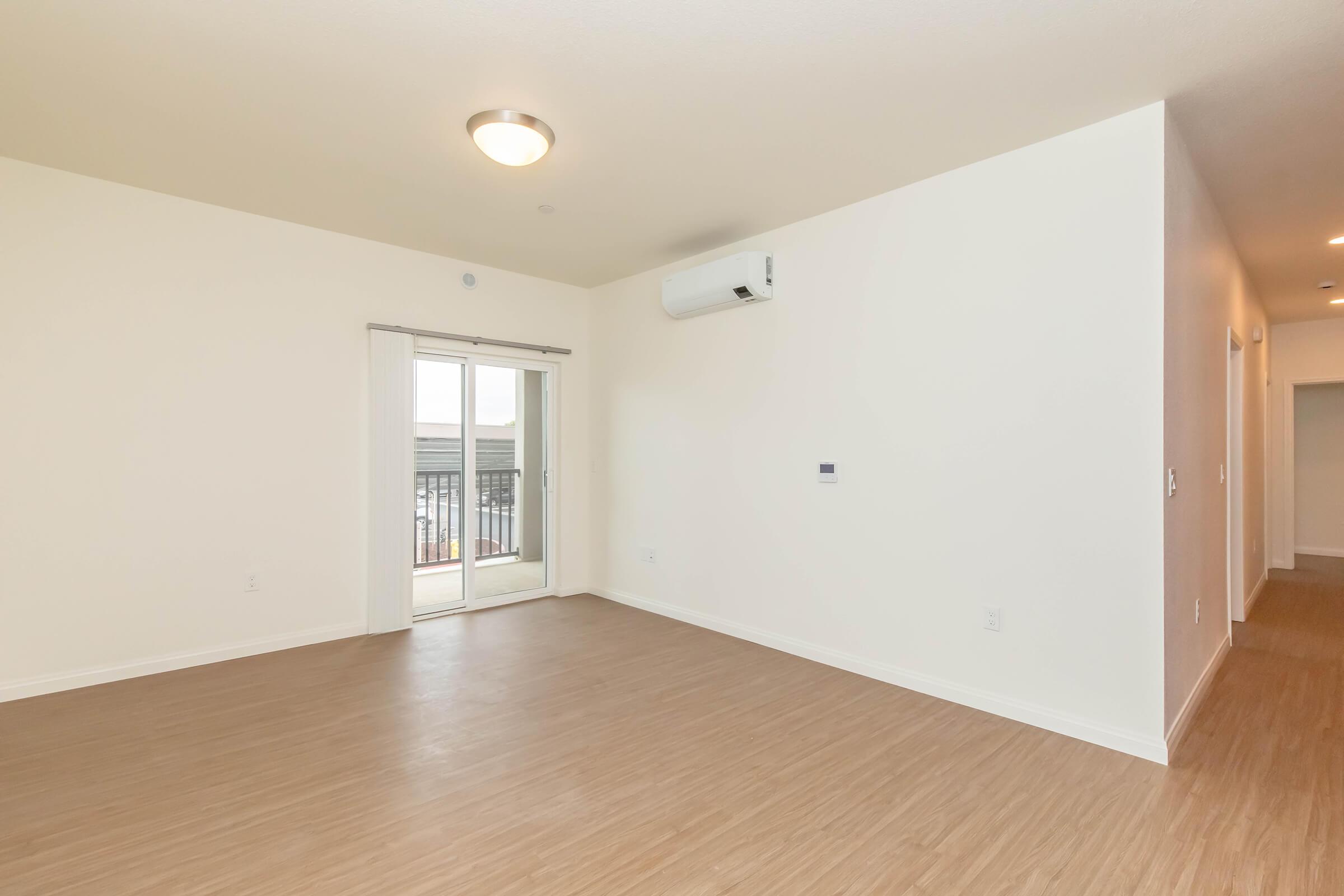 Empty room with light-colored walls and a seamless wooden floor. A sliding glass door leads to a small balcony, allowing natural light to enter. An air conditioning unit is mounted on the wall, and there are doorways leading to other rooms in the background.