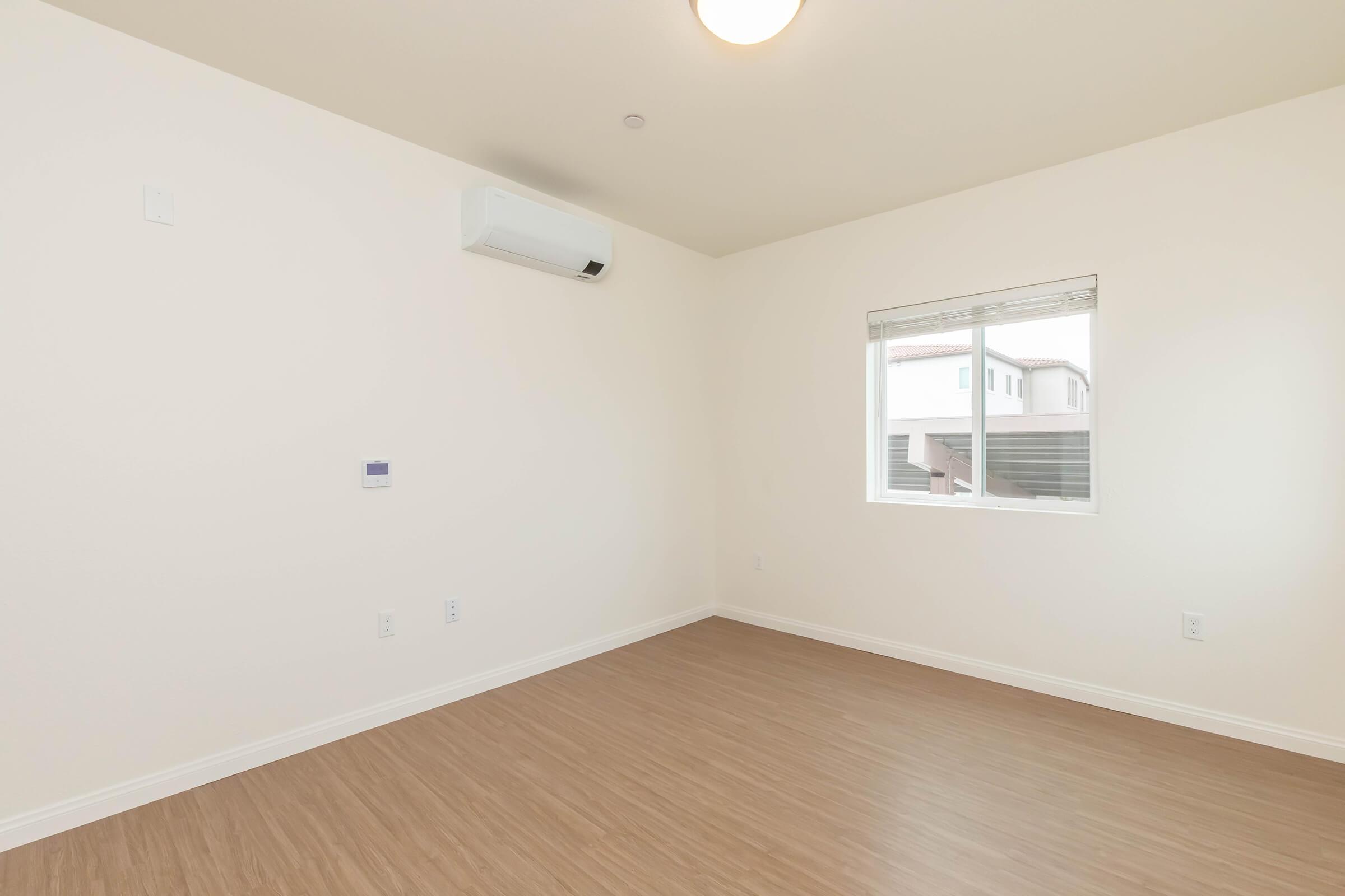 A sparsely furnished room with light-colored walls and a wooden floor. There is a window allowing natural light in, and an air conditioning unit mounted on the wall. The room appears empty, creating a minimalist and uncluttered atmosphere.