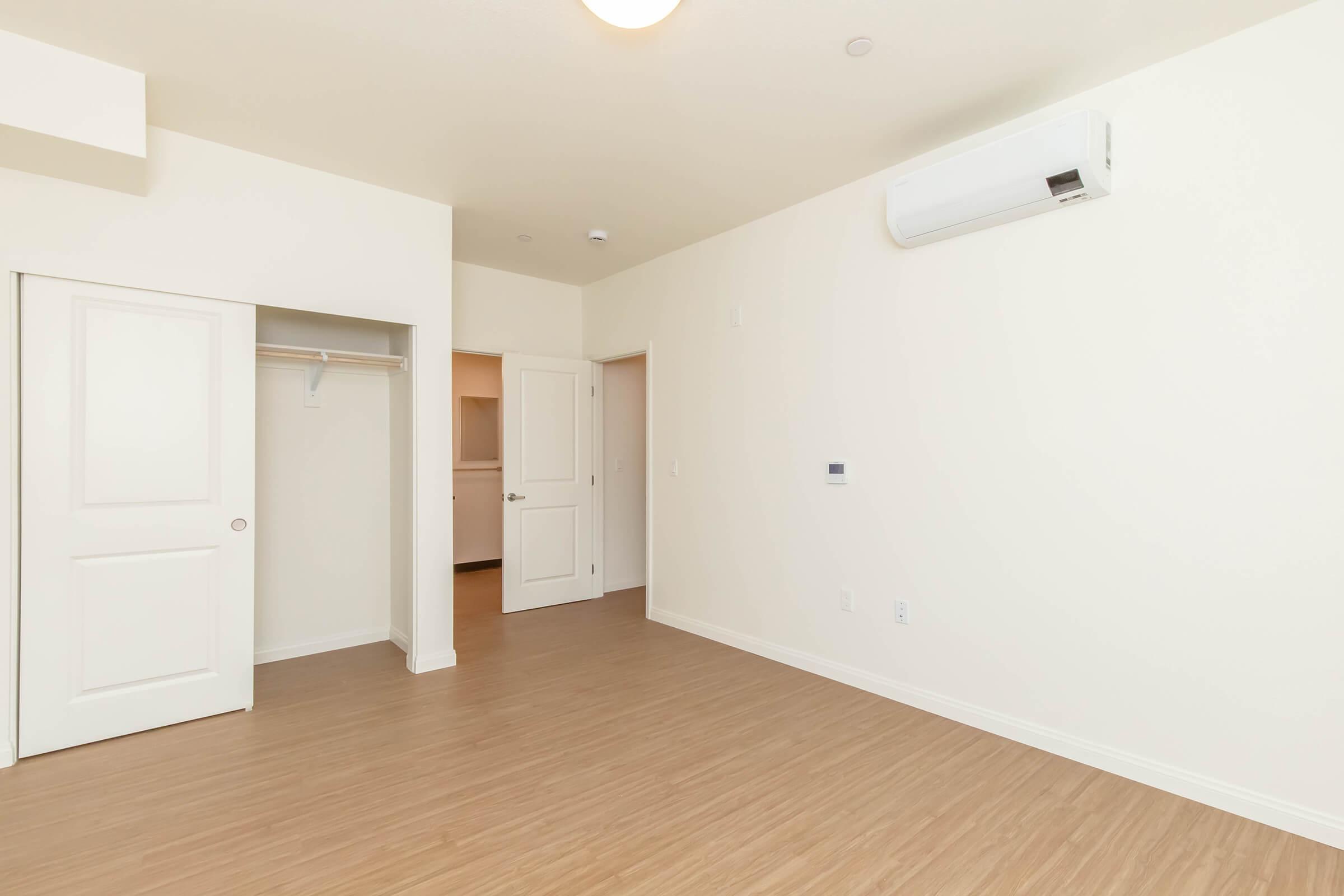 A vacant room with light-colored walls and a wooden floor. There is a closet with a sliding door on the left, and a doorway leading to another room in the background. An air conditioning unit is mounted on the wall, and the lighting is soft and bright. The space is clean and uncluttered.