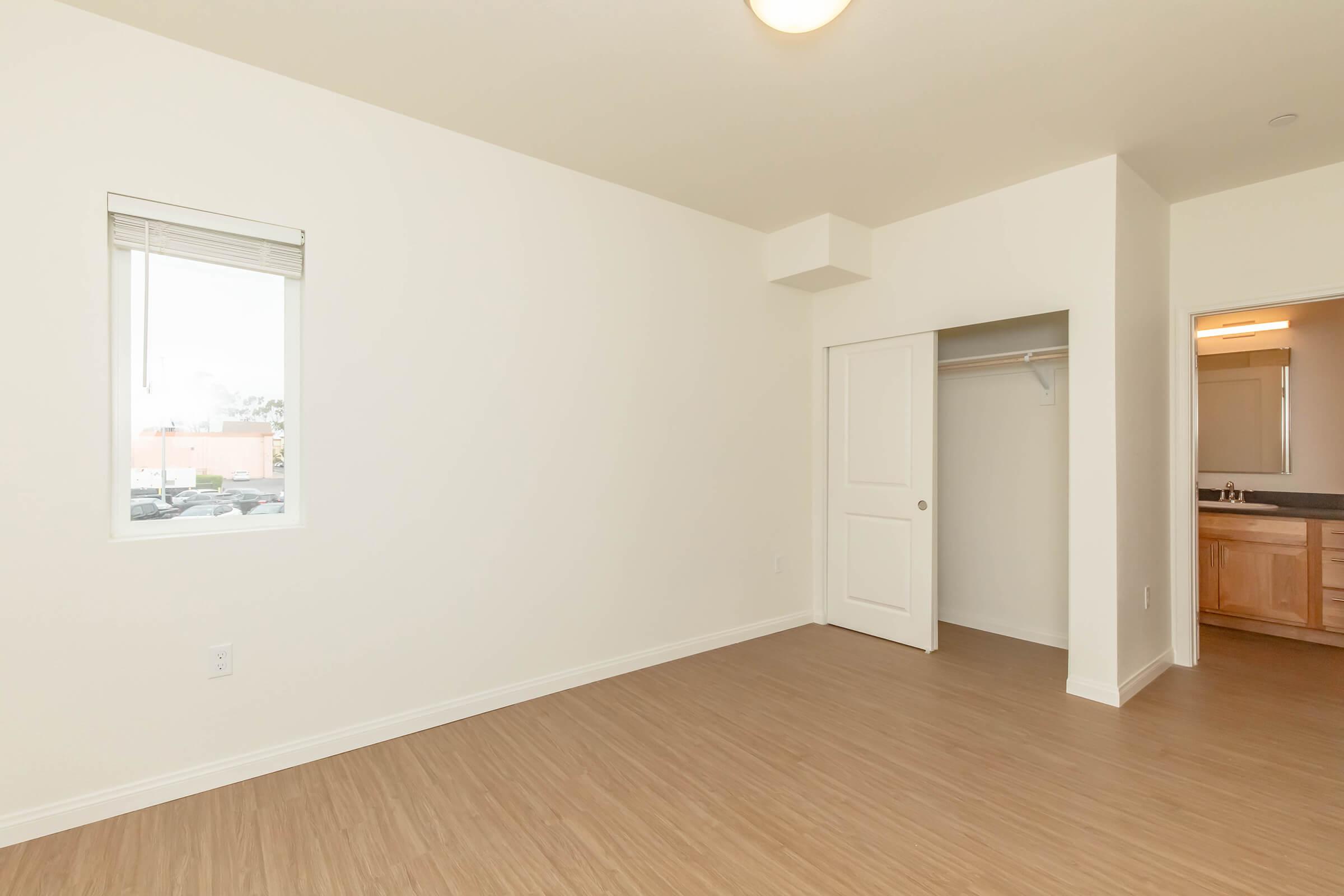 Empty room featuring light-colored walls and laminate flooring. A window allows natural light to enter, and there is a closet with a white door. To the right, a doorway leads to a bathroom with wooden cabinetry visible. The space is clean and minimalist, ideal for personal customization.