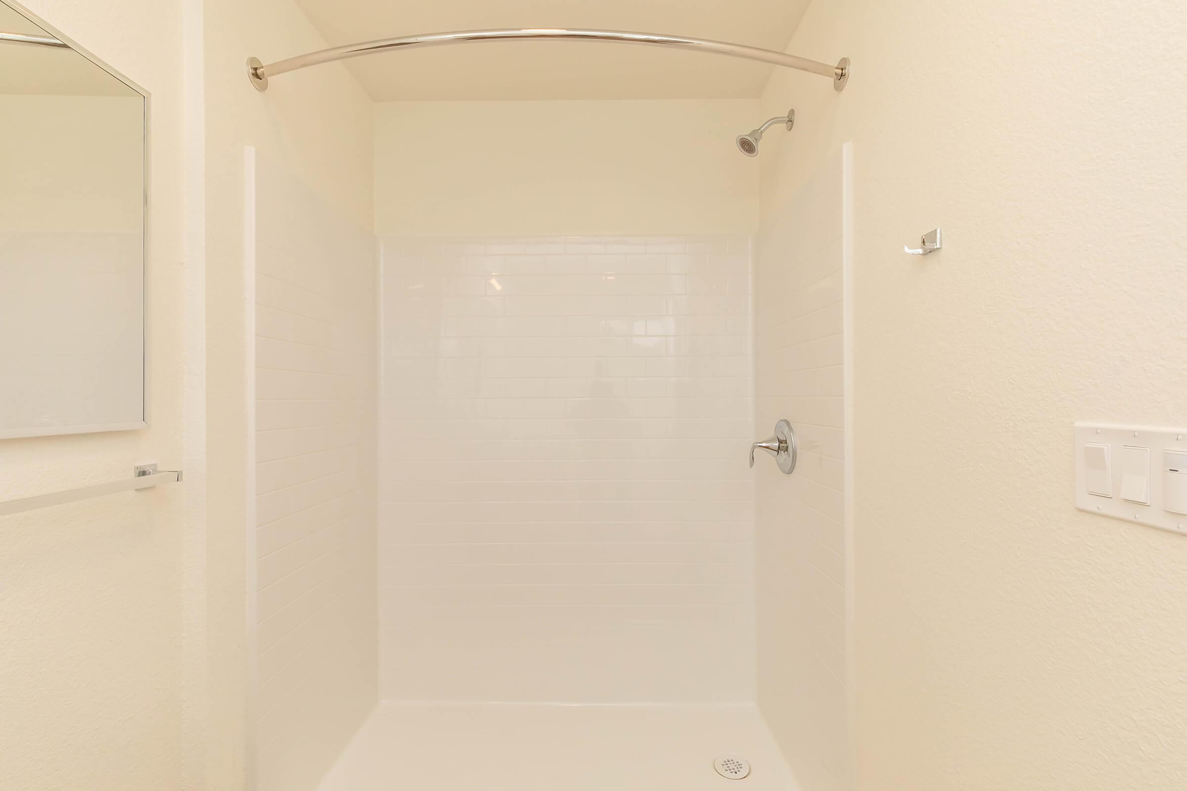 A clean, empty shower space with white tiled walls and a curved chrome shower rod. There are showerhead and wall-mounted faucet fixtures, and a small drain on the floor. A mirror is visible on the left side of the image. The overall aesthetic is bright and minimalistic with neutral colors.