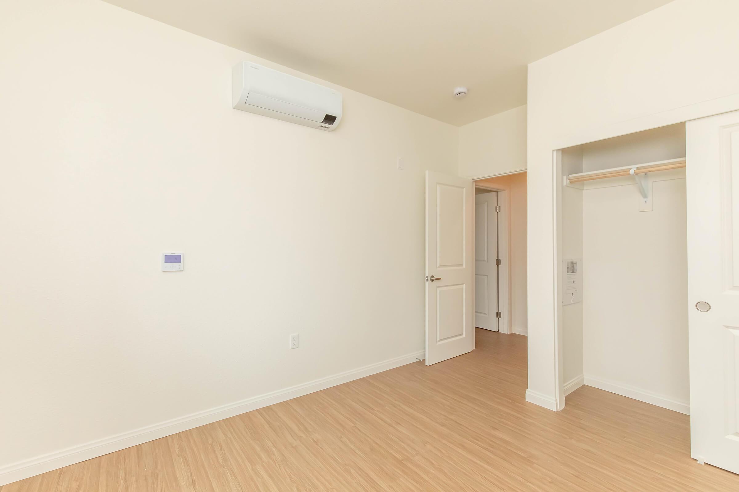 An empty room with light-colored walls and wooden flooring. There's an air conditioning unit mounted on the wall, a closed door leading to another space, and a closet with sliding doors. The room is well-lit, creating a spacious feel.