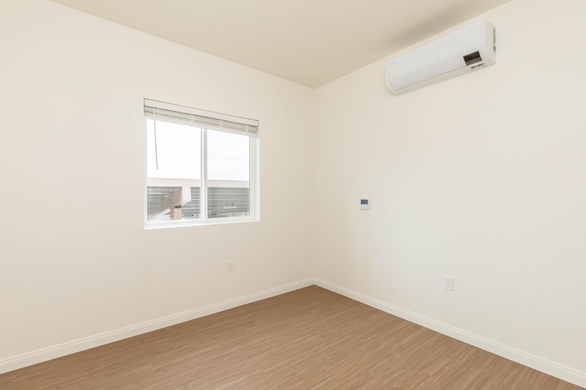 A minimalistic room with light-colored walls and a window. The flooring is a warm wooden finish, and an air conditioning unit is mounted on the wall. The space is empty, creating an airy and bright atmosphere.