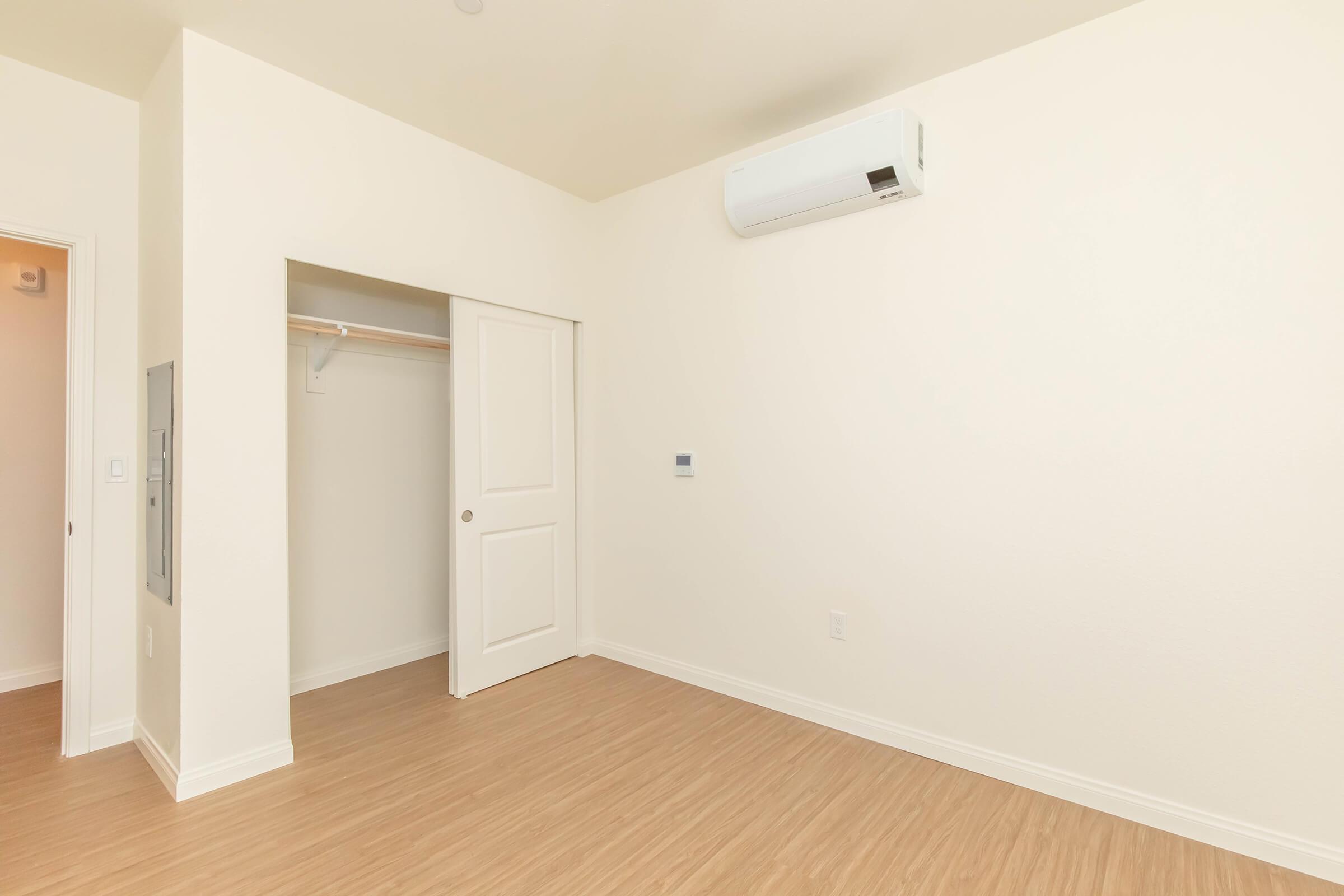 A clean, empty room with light-colored walls and wooden laminate flooring. There is a closet with an open door on one side and a white air conditioning unit mounted on the wall. The space appears bright and minimally furnished, creating an inviting atmosphere.