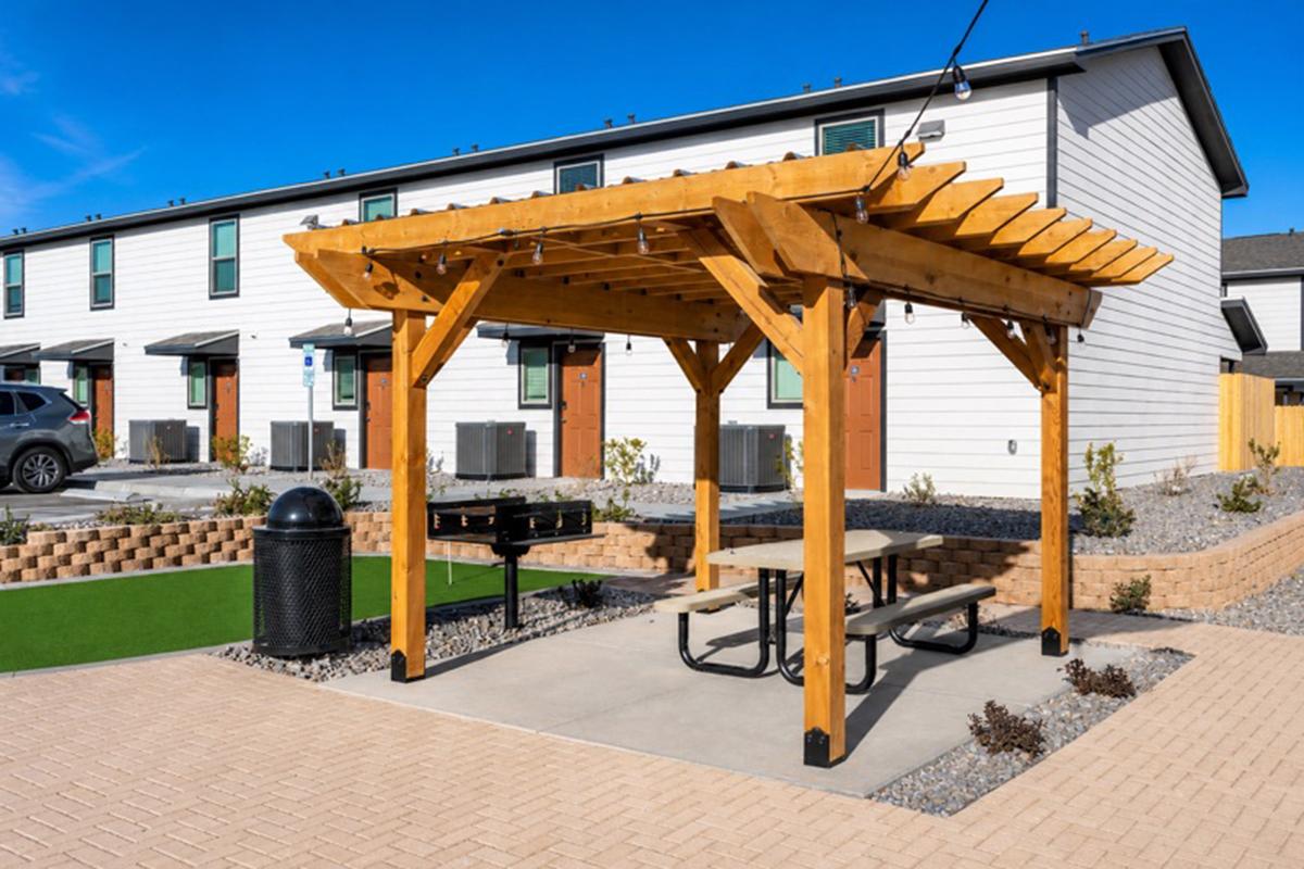 A wooden pergola with a picnic table and grill underneath, situated in a courtyard area. In the background, several modern residential buildings are visible. The ground is paved and has small shrubs, with a trash can nearby. The sky is clear and blue, indicating a sunny day.