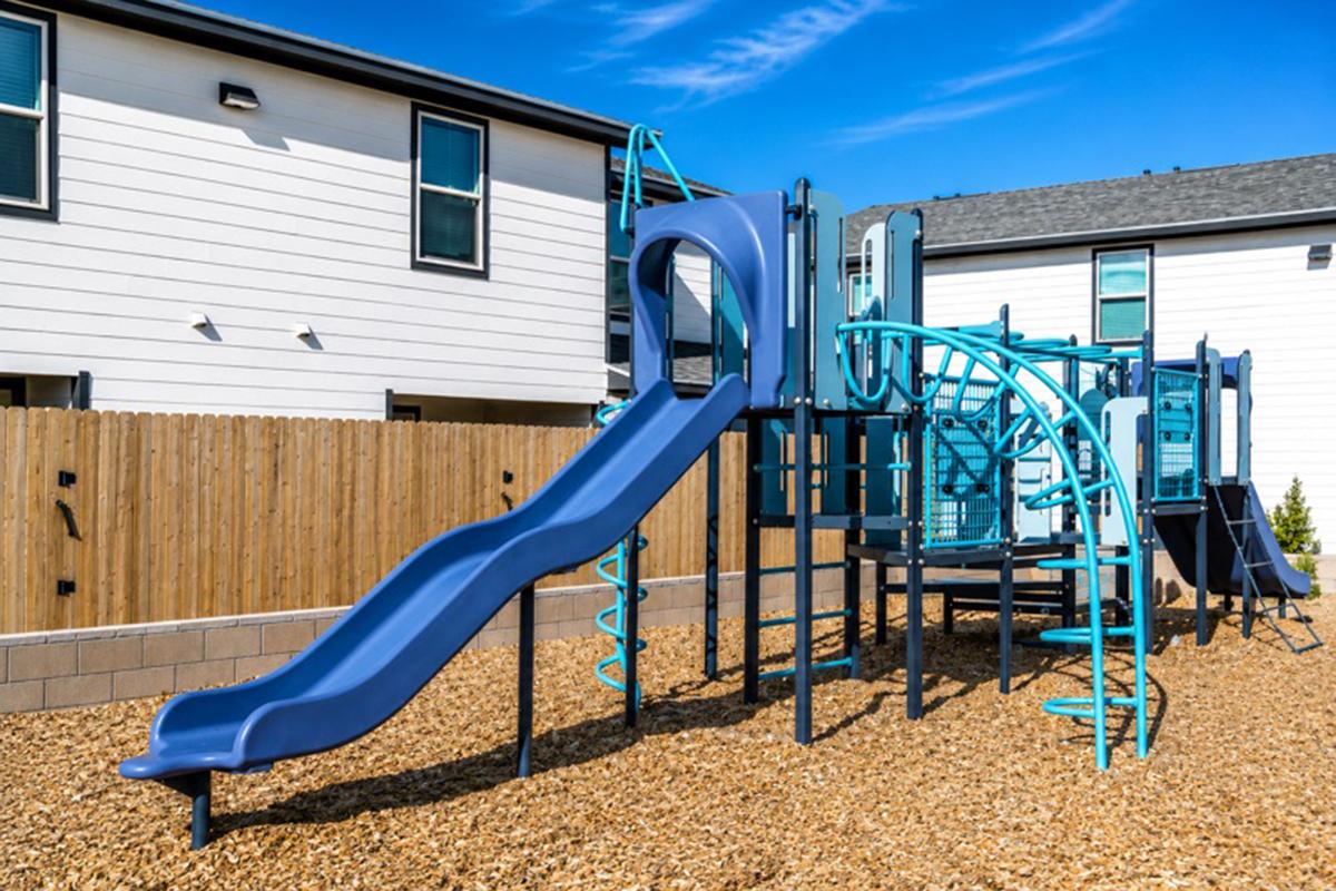 A modern playground featuring a blue slide, climbing structures, and various play equipment, situated on a bed of tan mulch. The background includes a wooden fence and residential buildings under a clear blue sky.